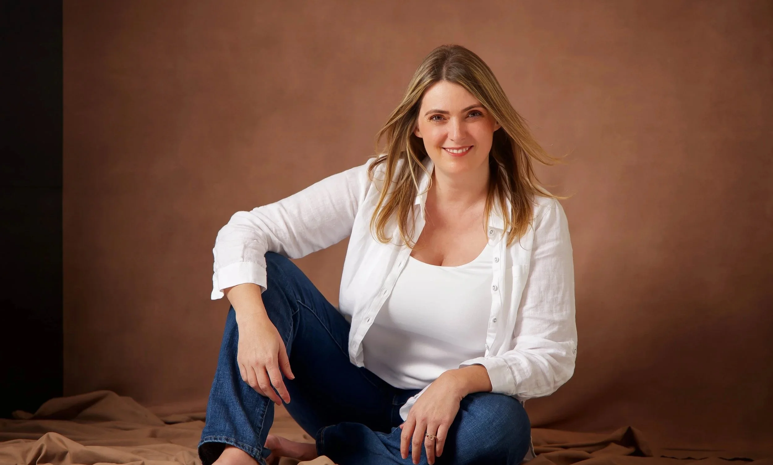 A woman with shoulder-length blonde hair sitting on a brown fabric backdrop, smiling at the camera, wearing a white shirt and blue jeans.