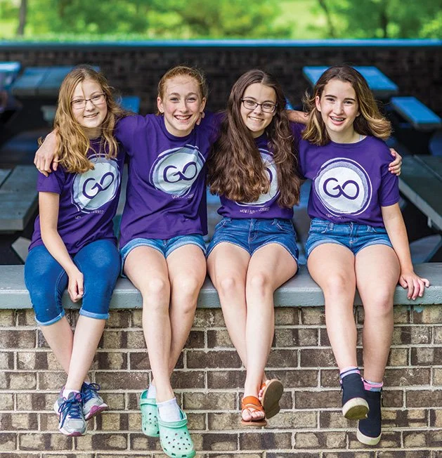 Four girls sitting side by side on a brick ledge outdoors, wearing matching purple T-shirts with a logo, denim shorts, and sneakers, smiling at the camera.