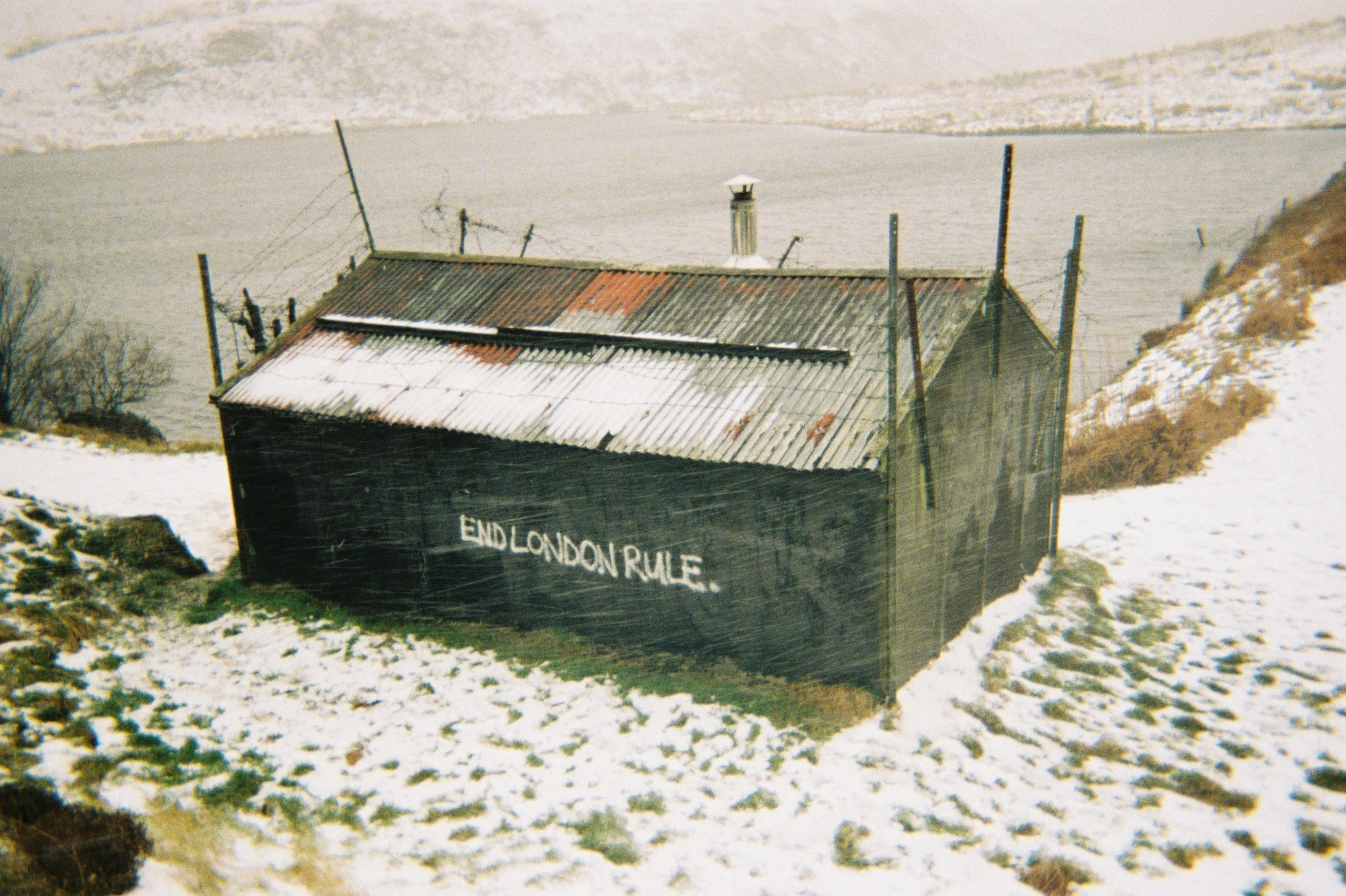 A black wooden shed on a snowy hillside with the words 'END LONDON RULE' painted on it, next to a body of water.