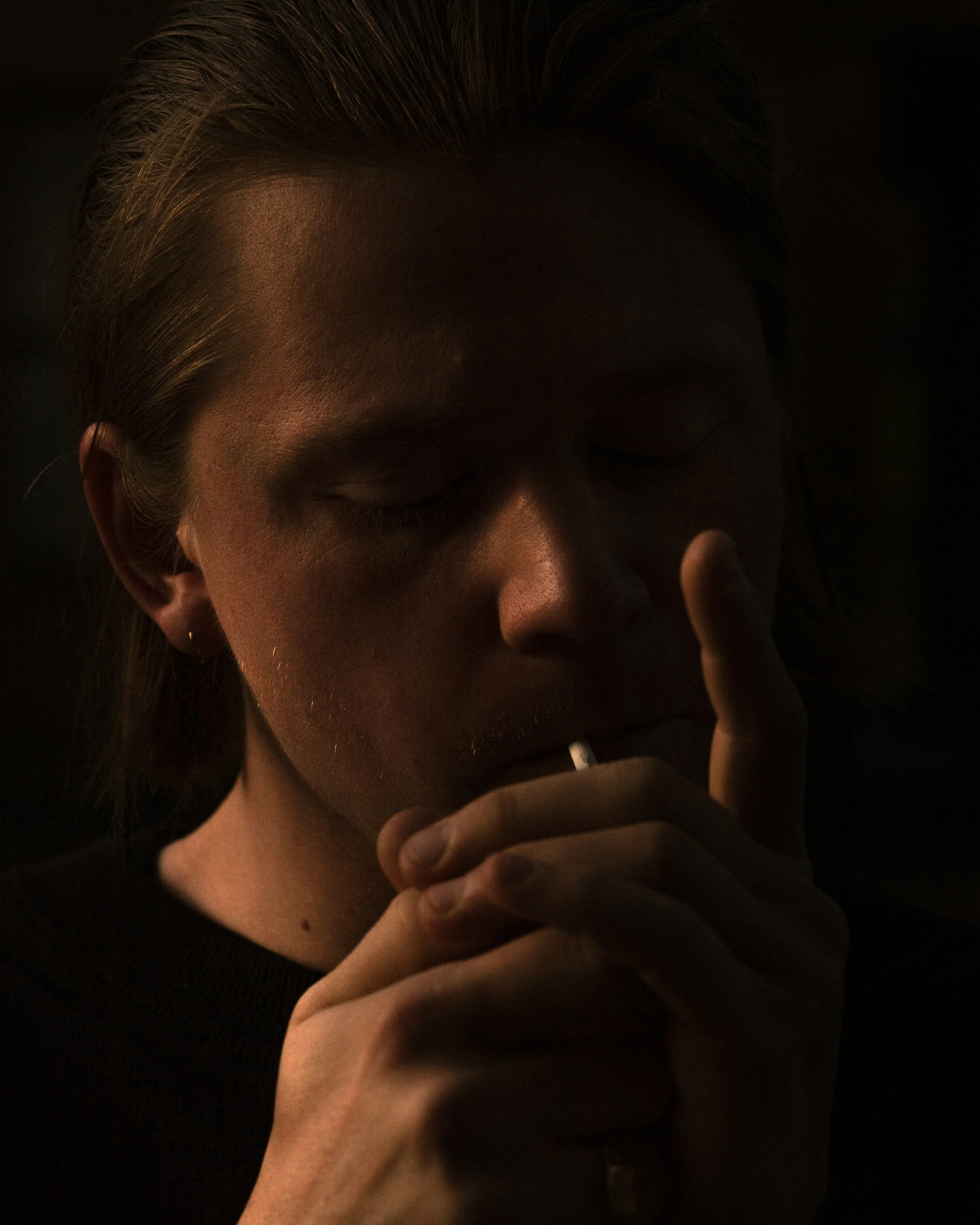 A person with closed eyes, lighting a cigarette with a match in a dark setting.