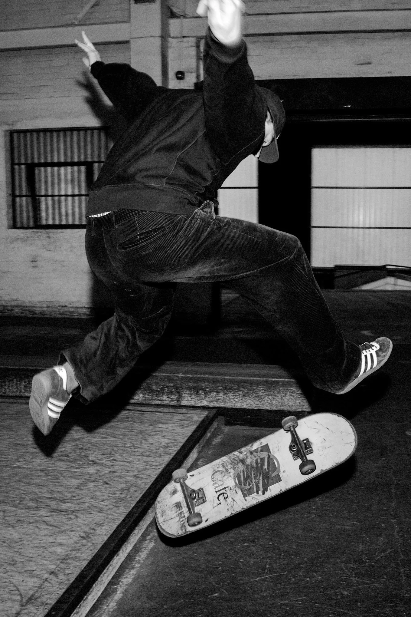 Skateboarder falls backward on a skate ramp indoors, with skateboard nearby, wearing jeans, sneakers, jacket, and cap.