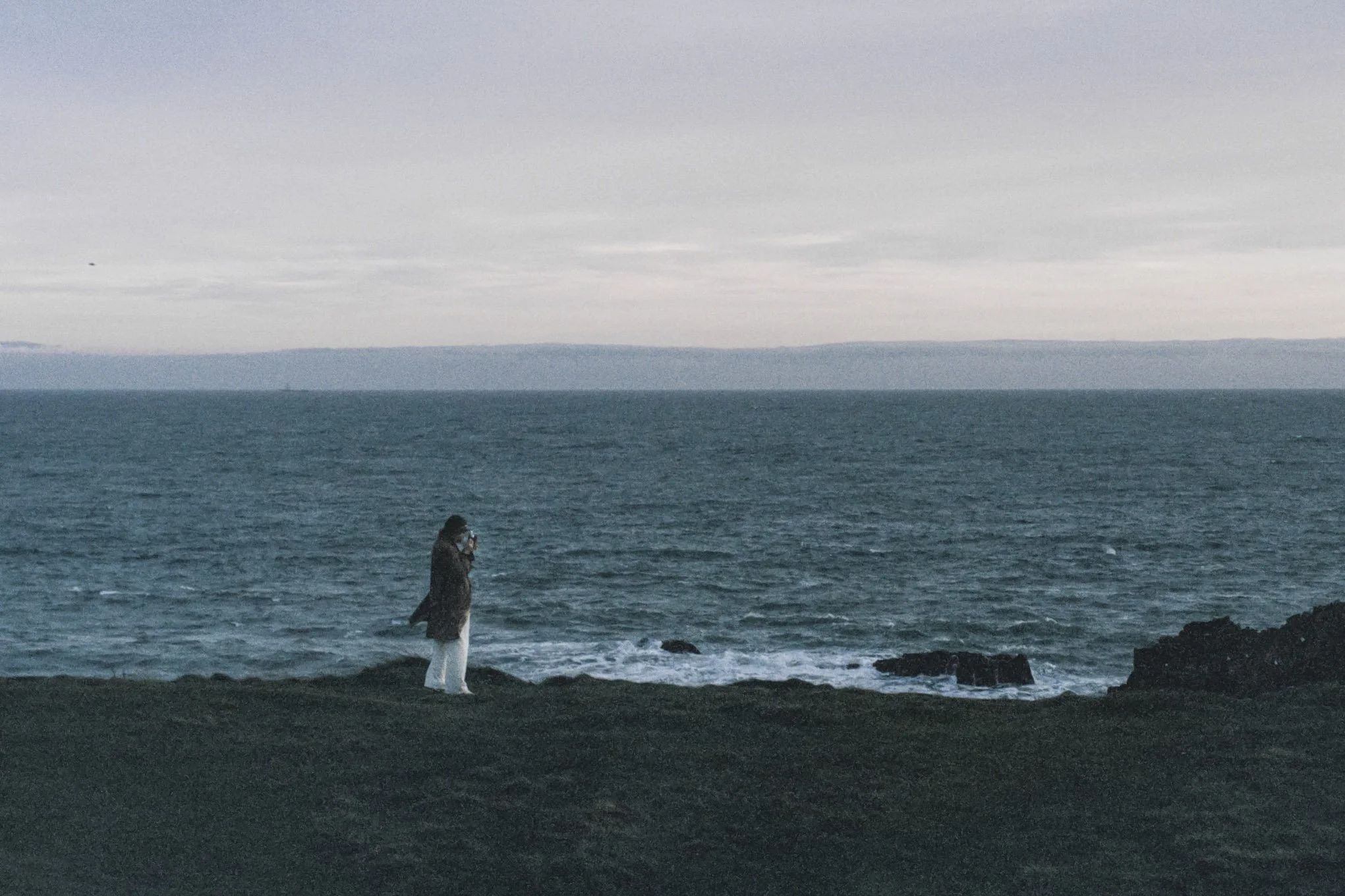 A person standing on the rocky shoreline looking at the ocean on a cloudy day.