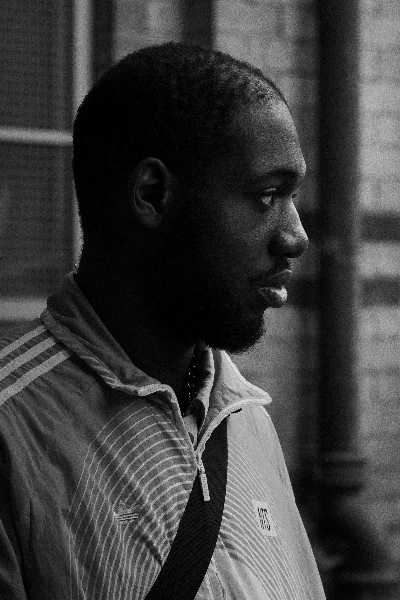Side profile of a man with short hair and beard, wearing a jacket with stripes and a logo, standing outdoors near a brick wall.