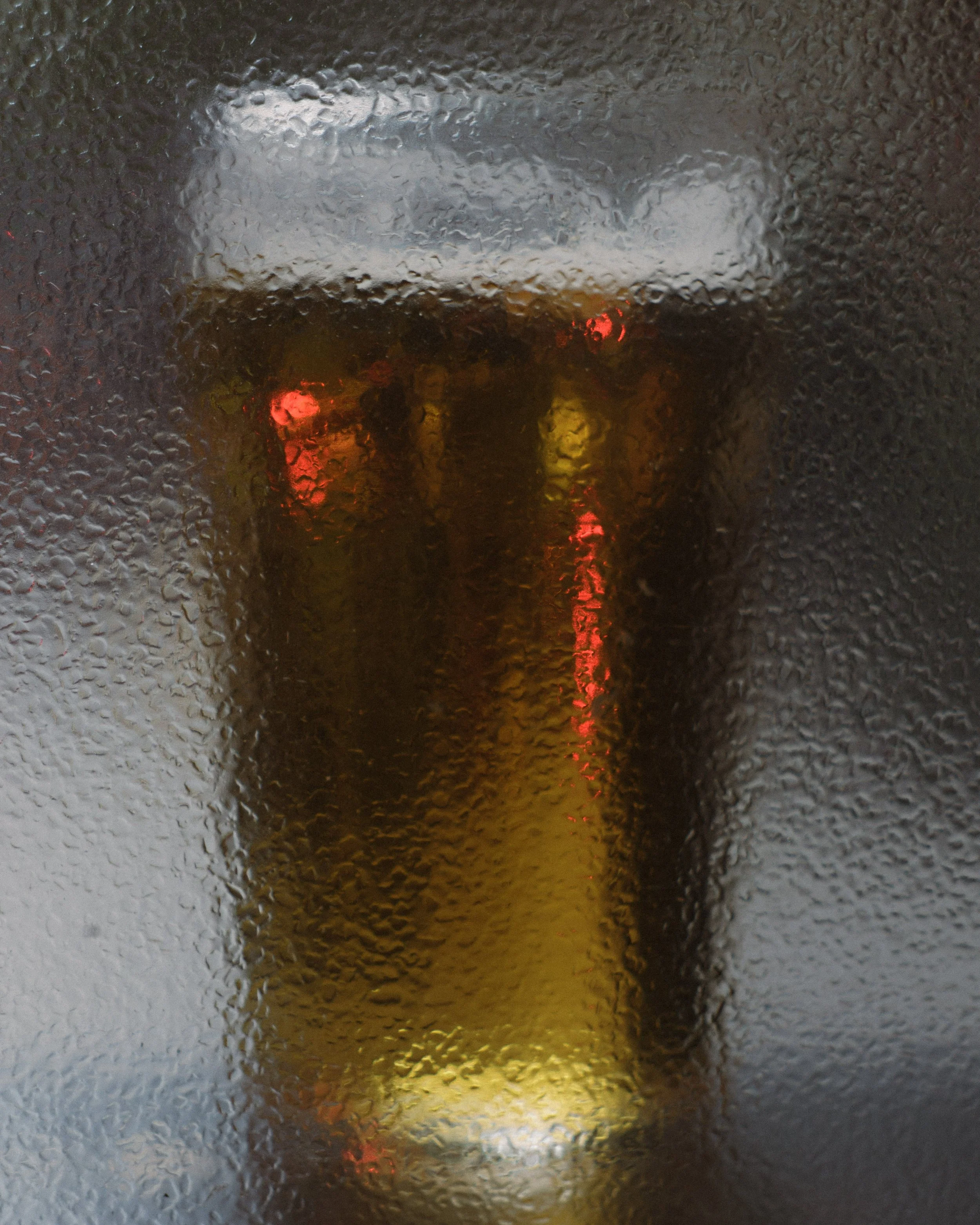 A glass of beer viewed through a rain-covered window, with blurred city lights in the background.