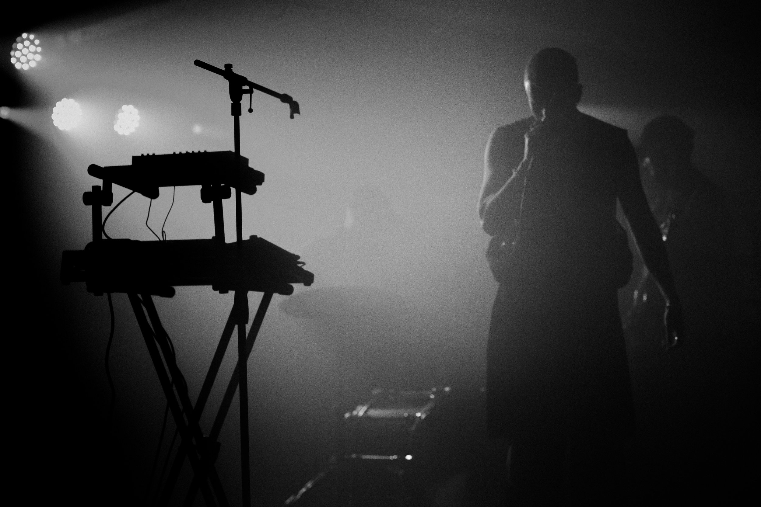 A musician holding a microphone on stage with a keyboard and microphone stand silhouetted in the foreground, foggy and with stage lights.
