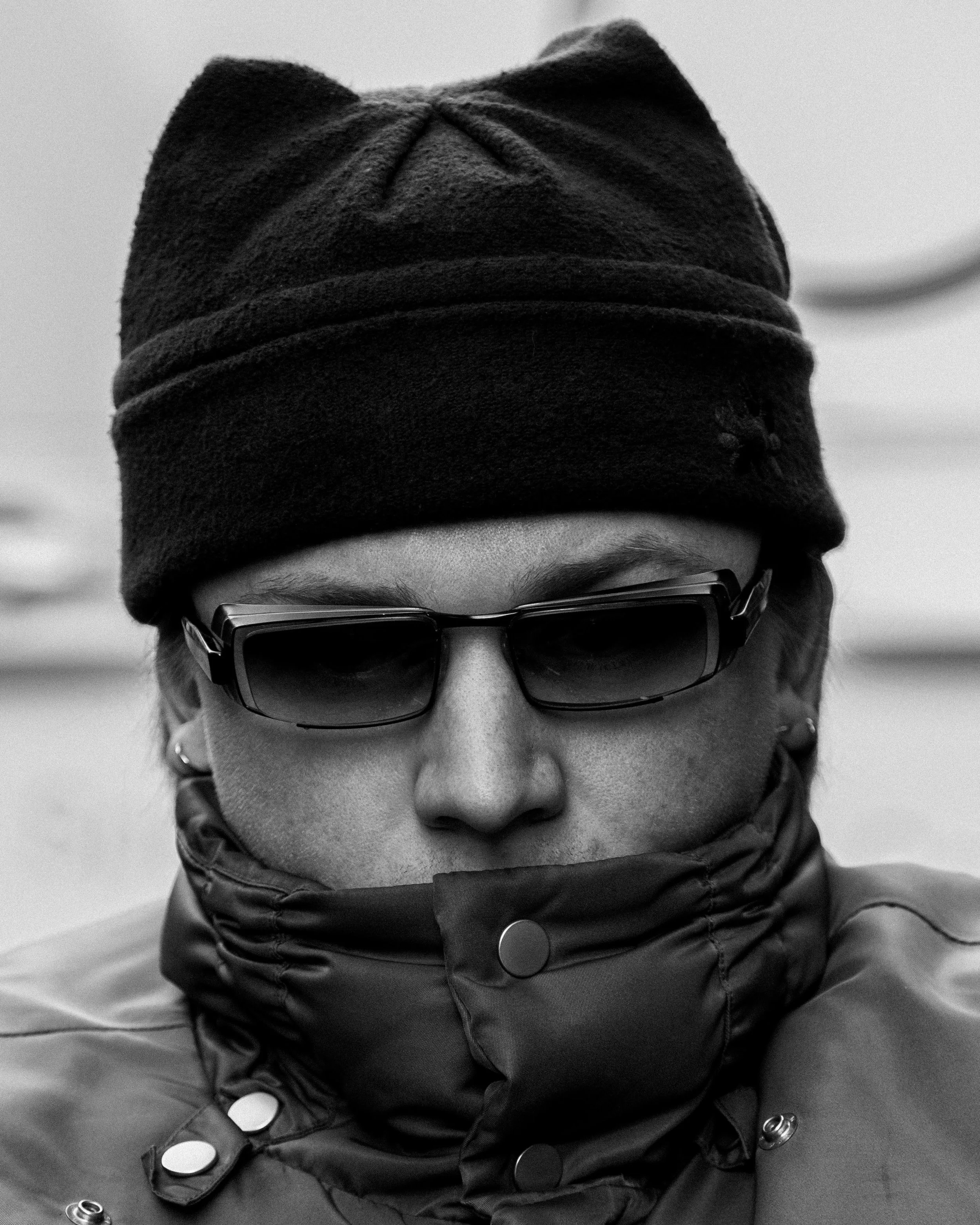 Close-up black and white photo of a person wearing a beanie, dark sunglasses, and a high-collared jacket.