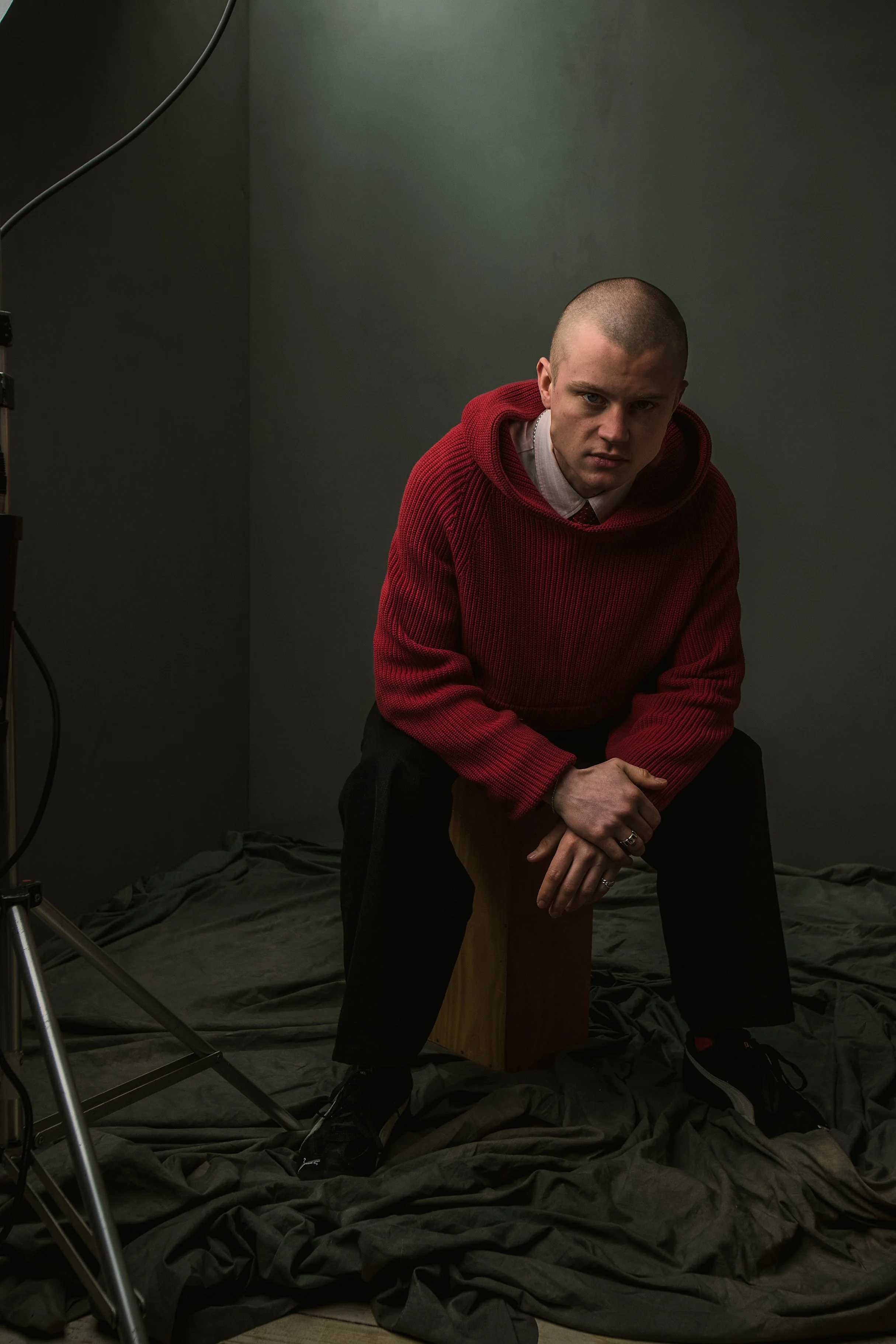 Young man in a red hoodie sitting on a wooden block with a dark backdrop, surrounded by studio equipment and black fabric on the floor.