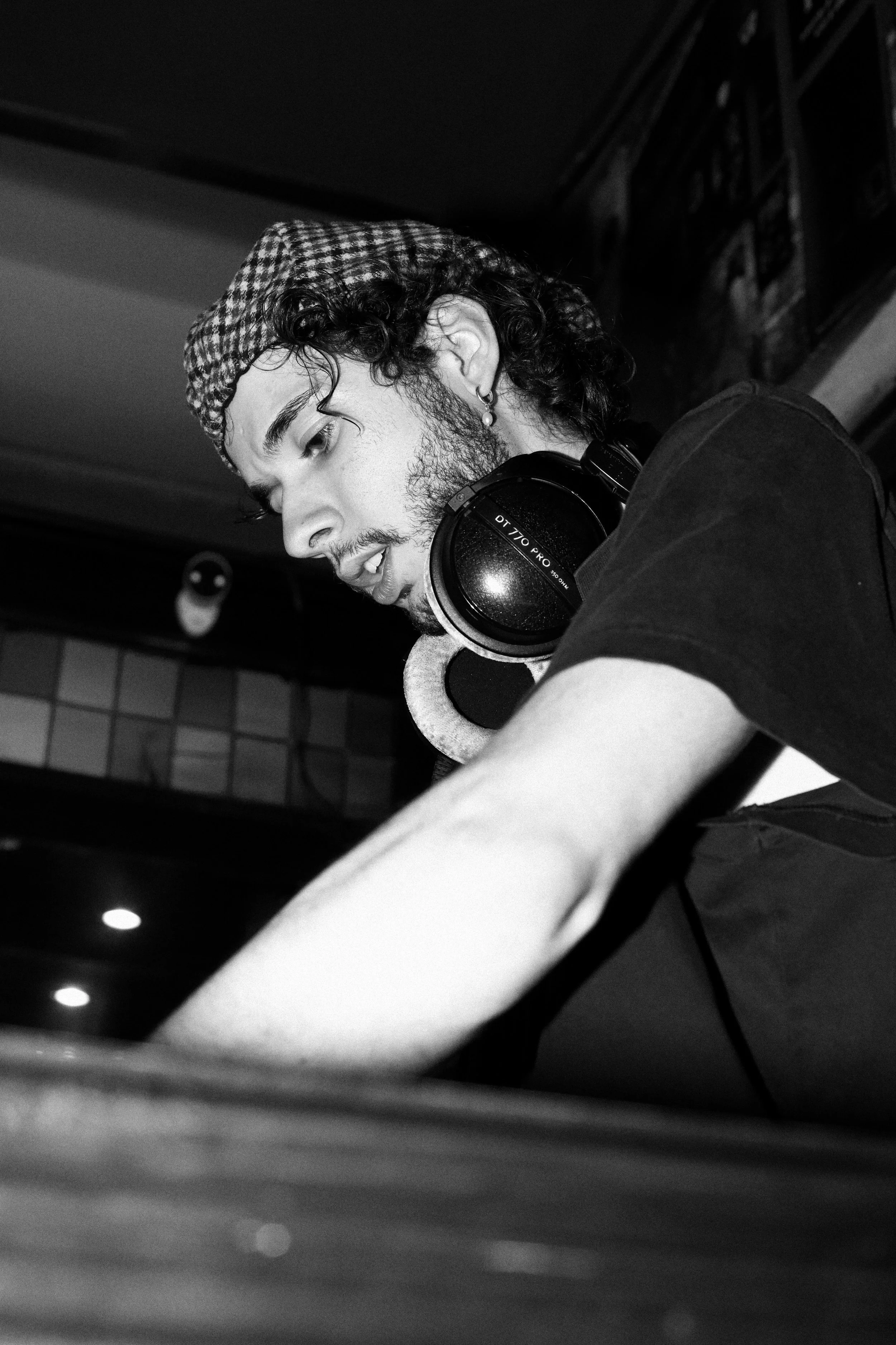 A young man with curly hair, wearing a checkered cap, is focused on something in front of him. He has headphones around his neck and is wearing a dark t-shirt. The setting appears to be indoors, possibly a cafe or studio, with shelves or artwork in t
