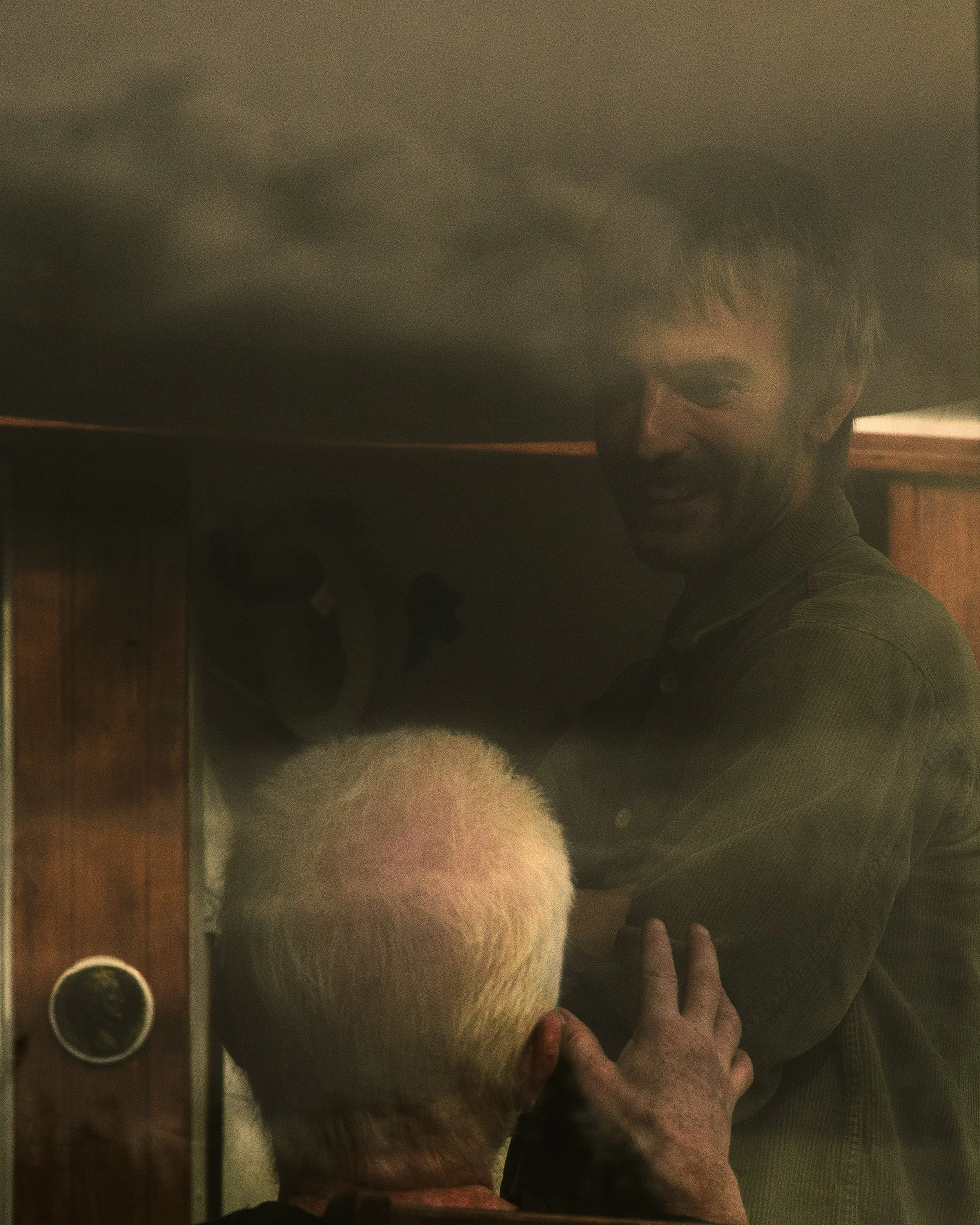 Two men are engaged in a friendly conversation; one elderly with white hair and the other younger with dark hair, smiling, with wooden walls in the background.