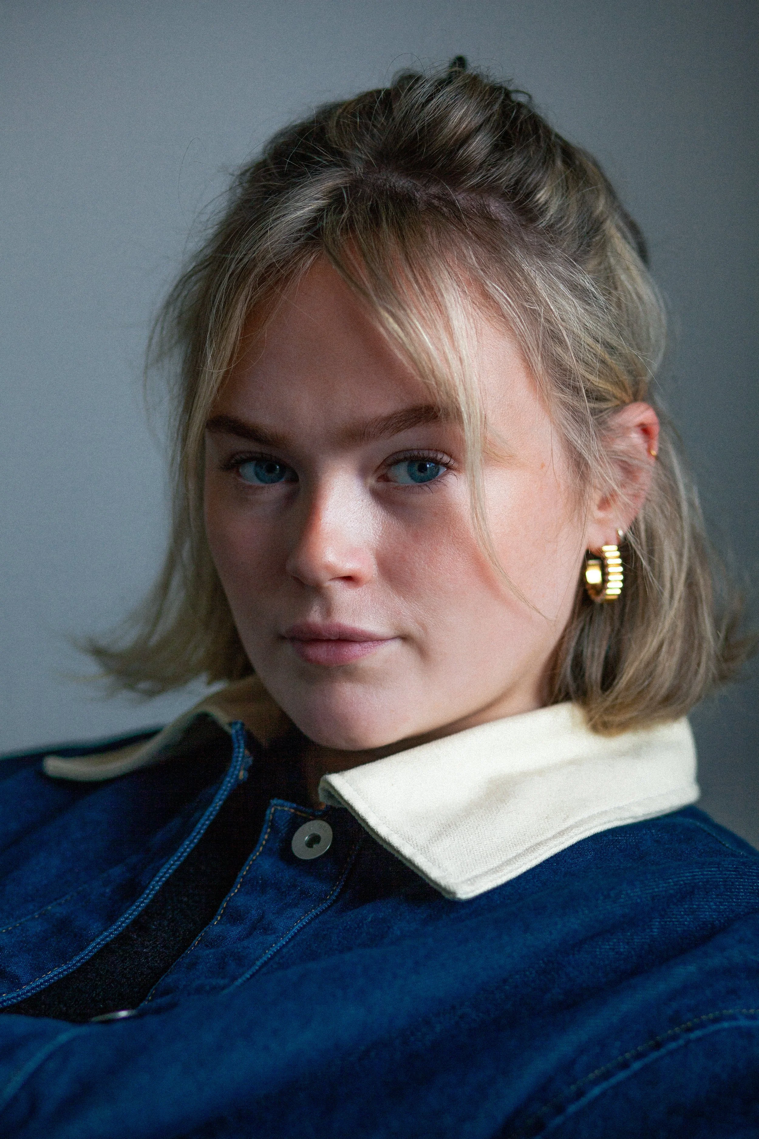 Close-up of a young woman with blond hair, blue eyes, wearing gold hoop earrings, a denim jacket with a white collar, set against a plain background.