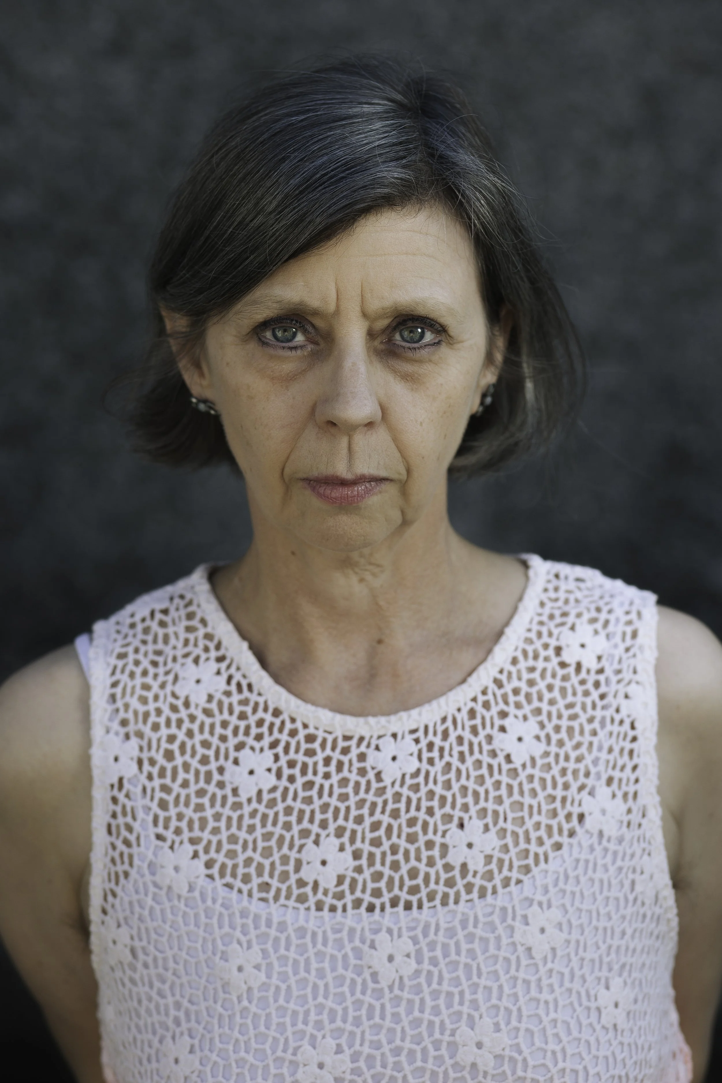 A woman with short dark hair and blue eyes wearing a white crochet top, standing against a dark gray background.