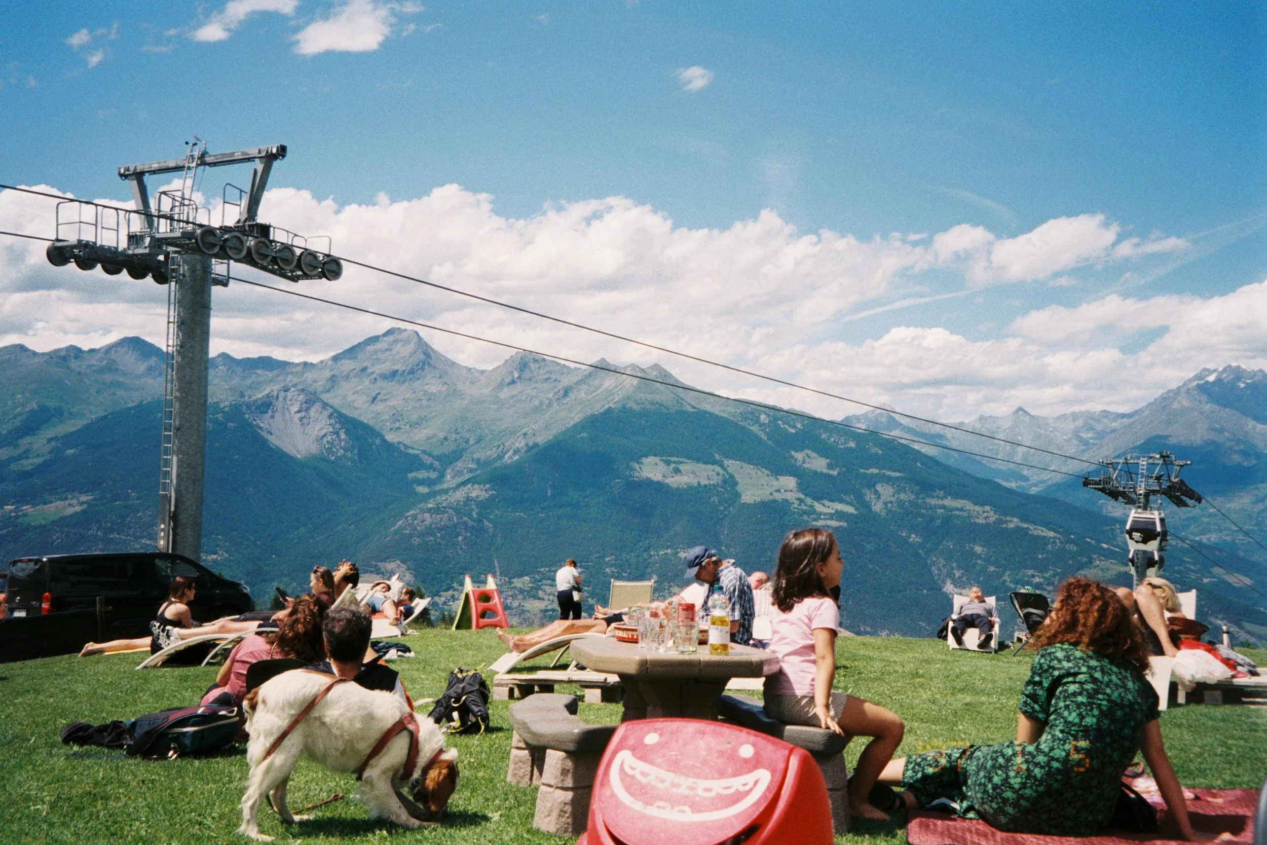 People relaxing on a grassy hill with mountain scenery, some lying on lounge chairs, others sitting or standing, with a dog nearby; ski lift towers and cables in the background, under a partly cloudy sky.