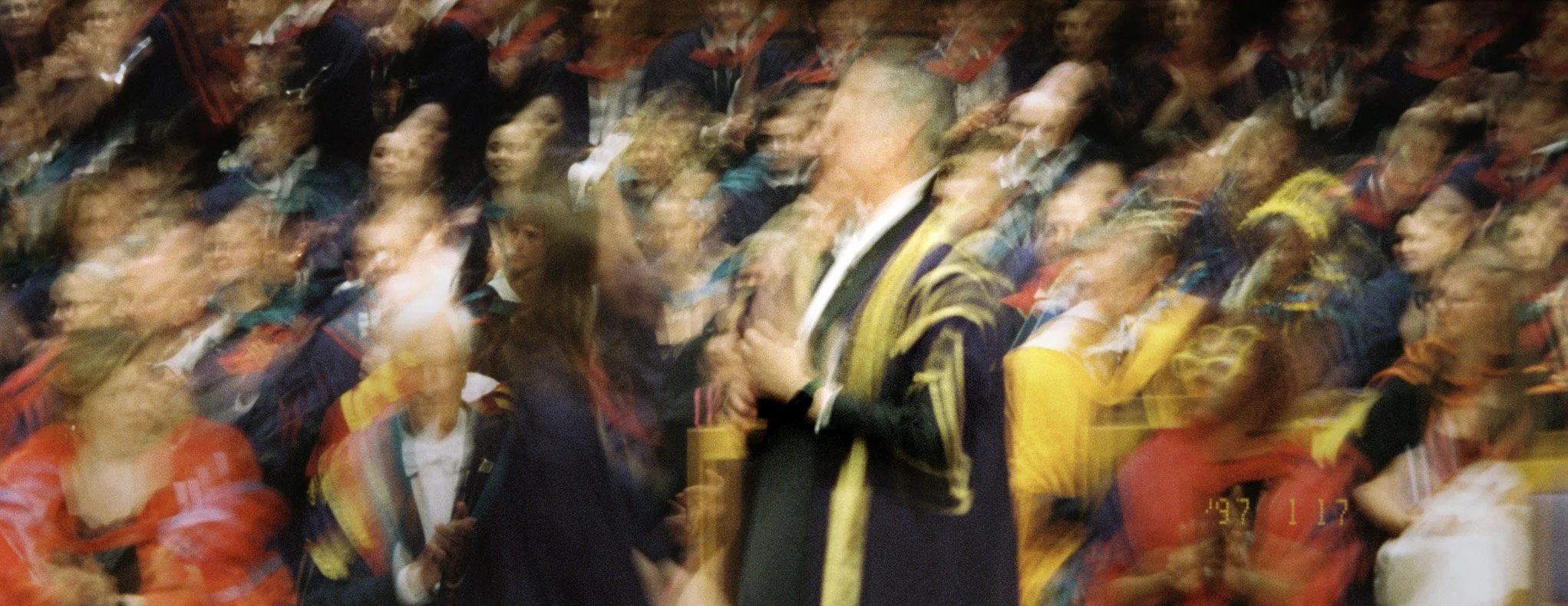 Blurry photo of a graduation ceremony with students in caps and gowns and a faculty member in academic regalia, shaking hands.