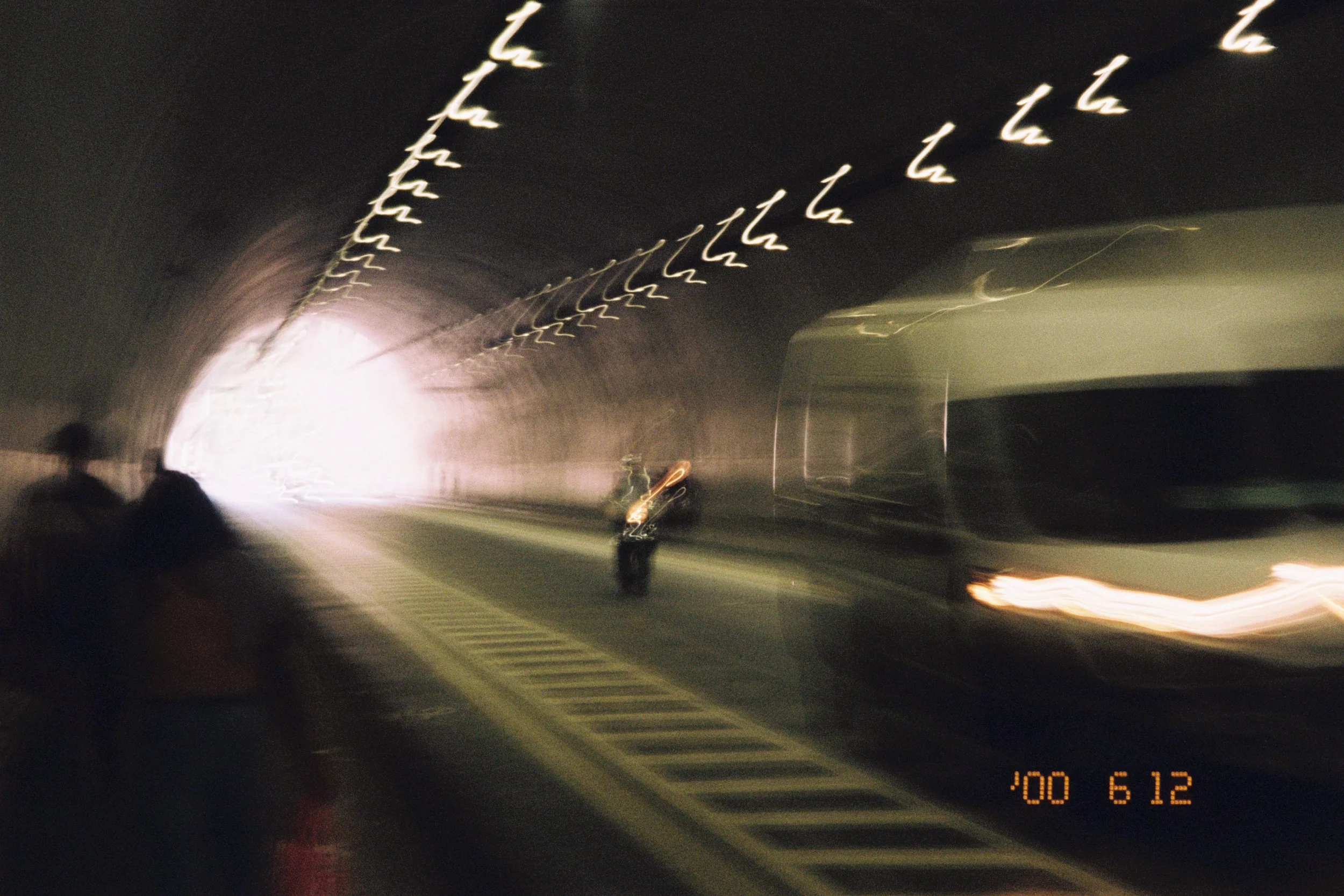 Blurred photo of a tunnel with streaks of light, two people sitting on the left side, and a bus on the right side, with a motorcycle in the middle, all under bright lights near the tunnel's opening.