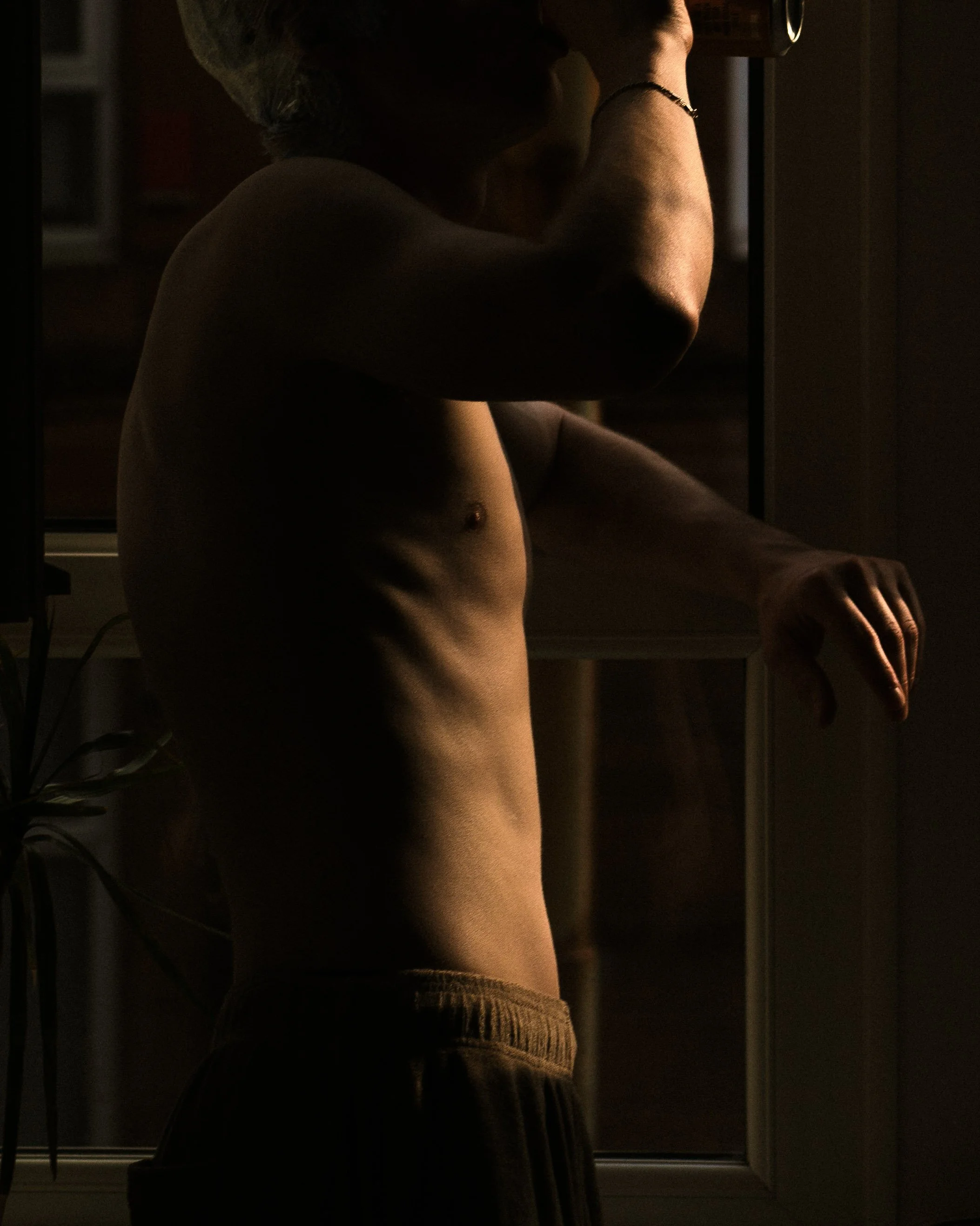 A shirtless man standing near a window, illuminated by warm, low lighting.