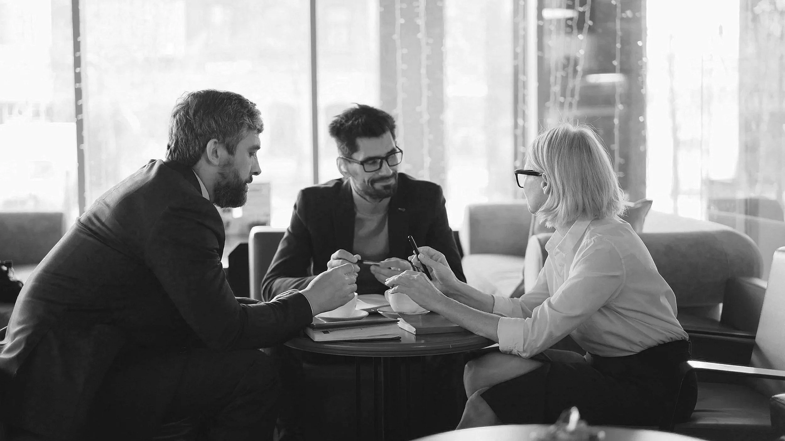 Three people in a business meeting sitting around a table, engaged in conversation, in a modern office with large windows.