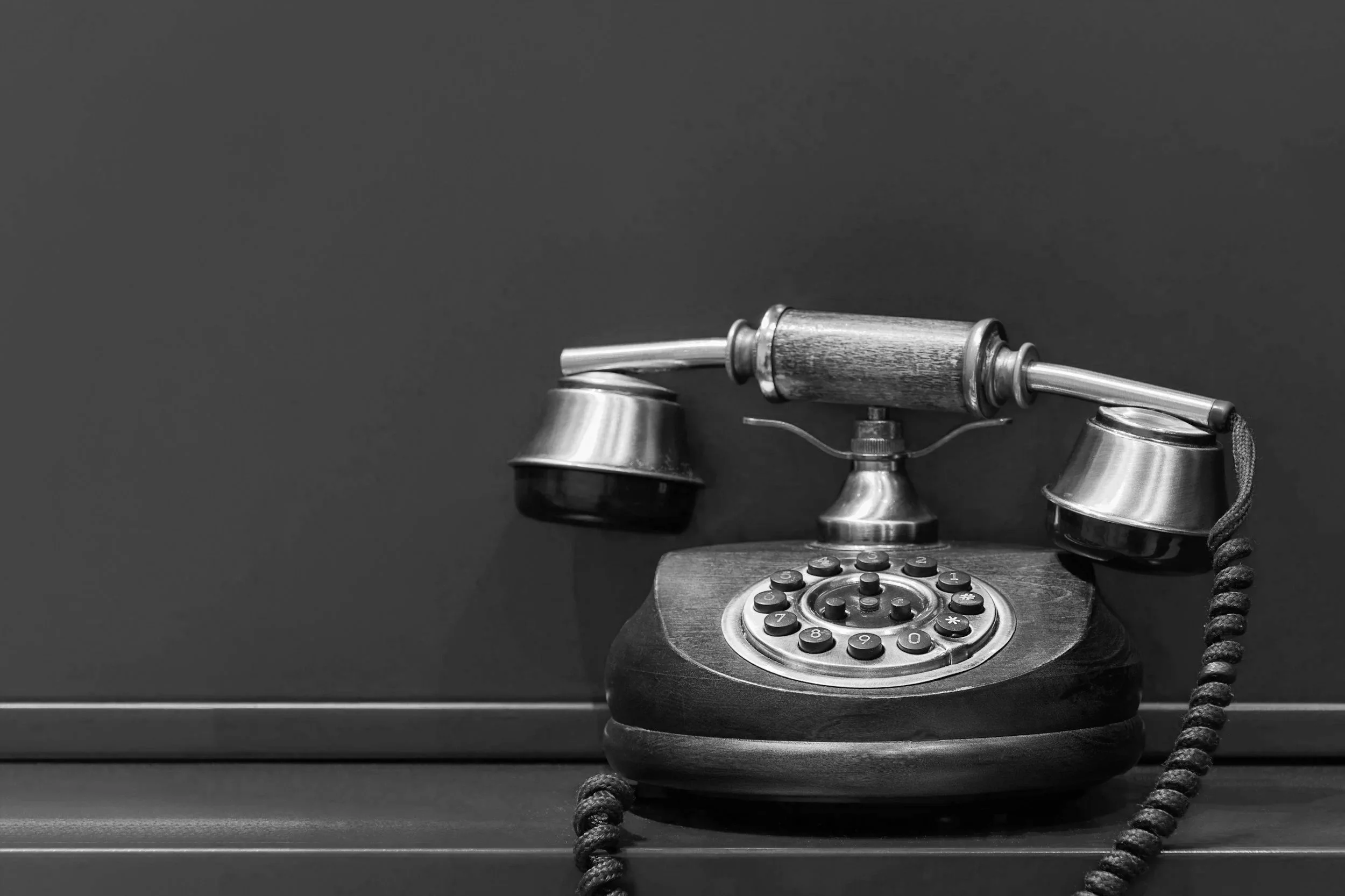 Black and white photo of an vintage rotary telephone with a wooden handle, placed on a dark desk against a plain background.