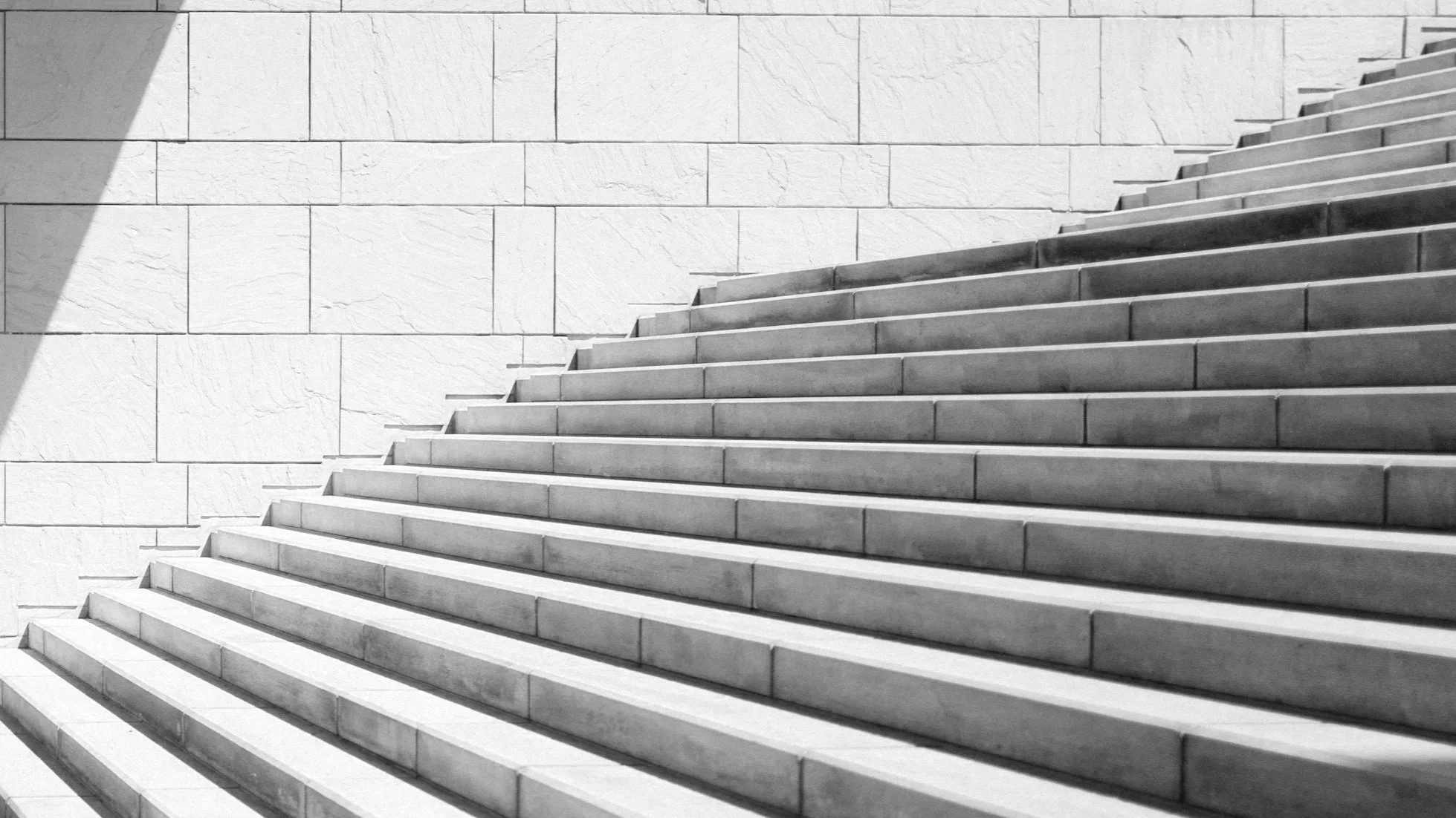Concrete stairs ascending alongside a large stone wall, with a shadow cast on the upper left corner.