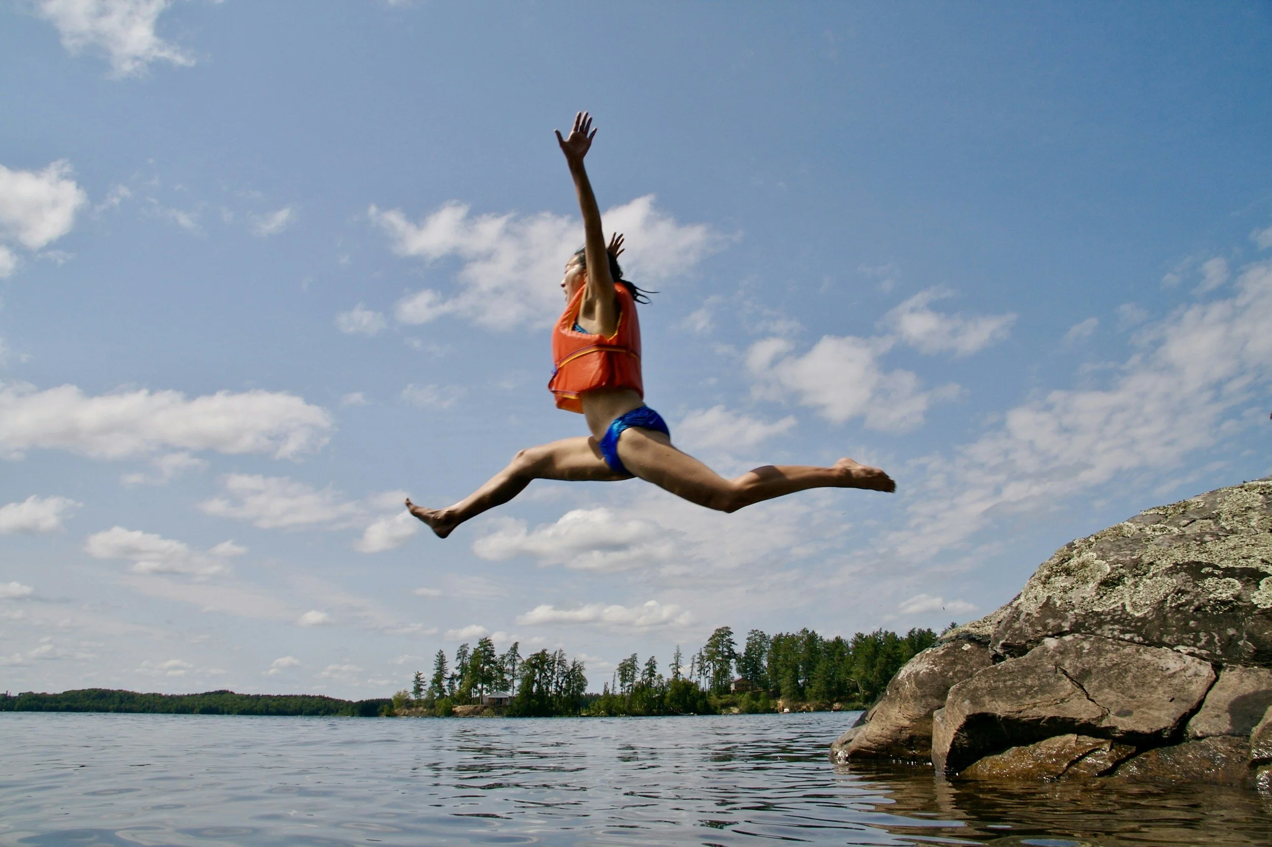 A person wearing a blue bikini and an orange life vest jumps from a pile of rocks into a lake. Her arms and legs are flung wide and she looks to be joyfully shouting.