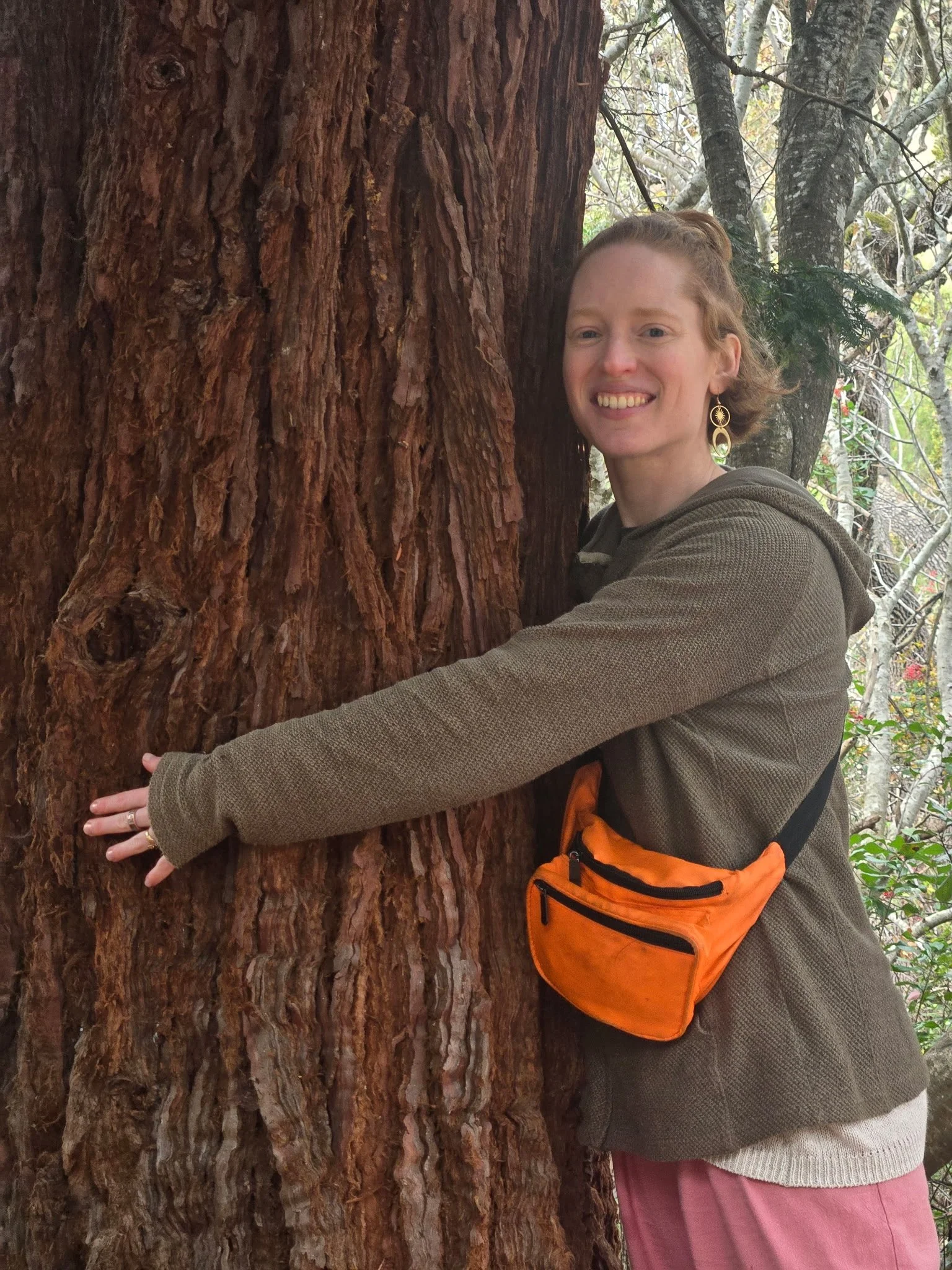 A photo of Serra Shelton PT, DPT. She is wrapping her arms around the trunk of a large redwood tree and smiling.
