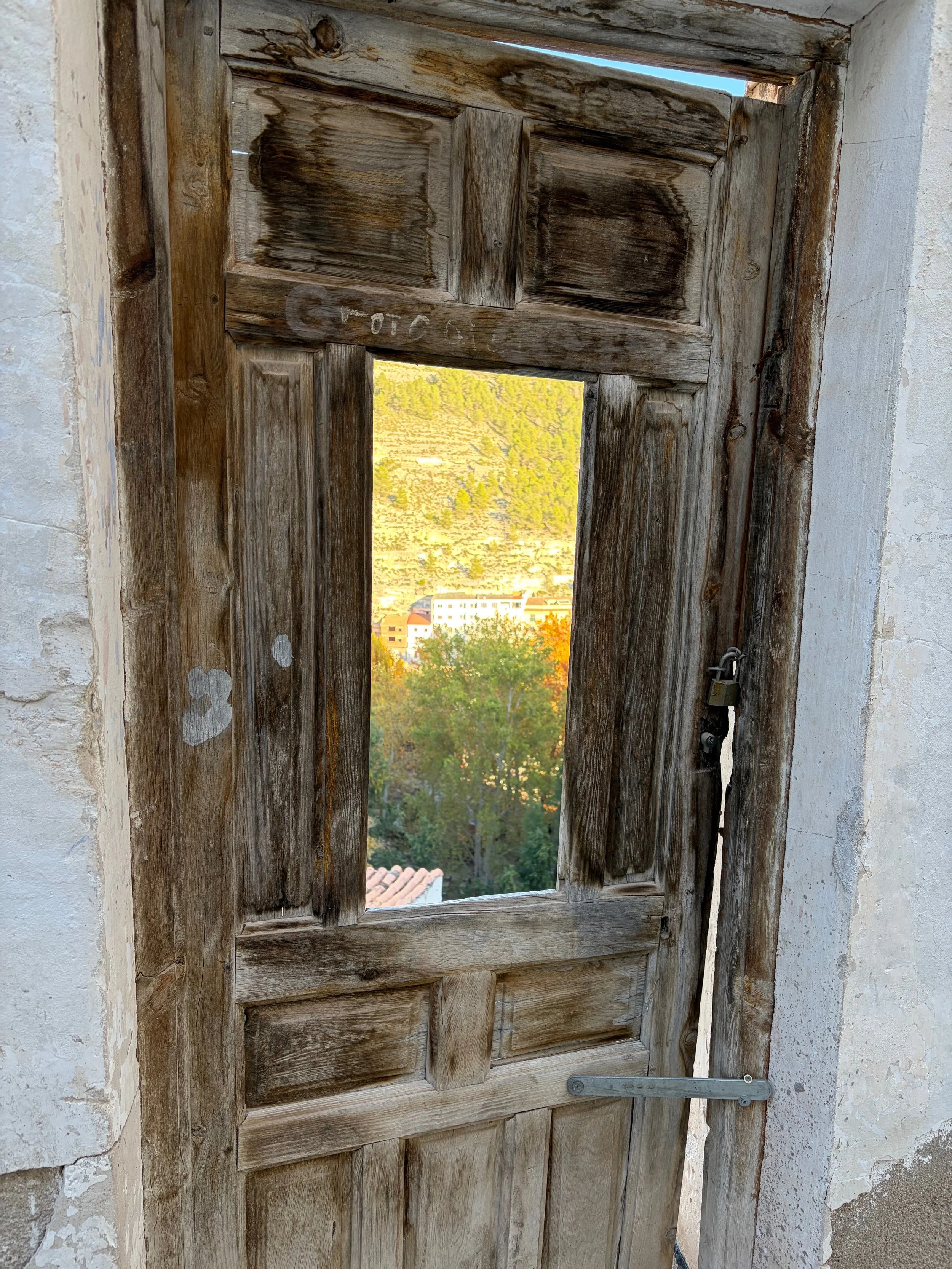 Old wooden window with a small opening, locked with a metal bar, overlooking a landscape of trees and buildings in the distance. Photo taken Spain, 2025. Doors are a metaphorical for curiosity, being open, and being grounded.