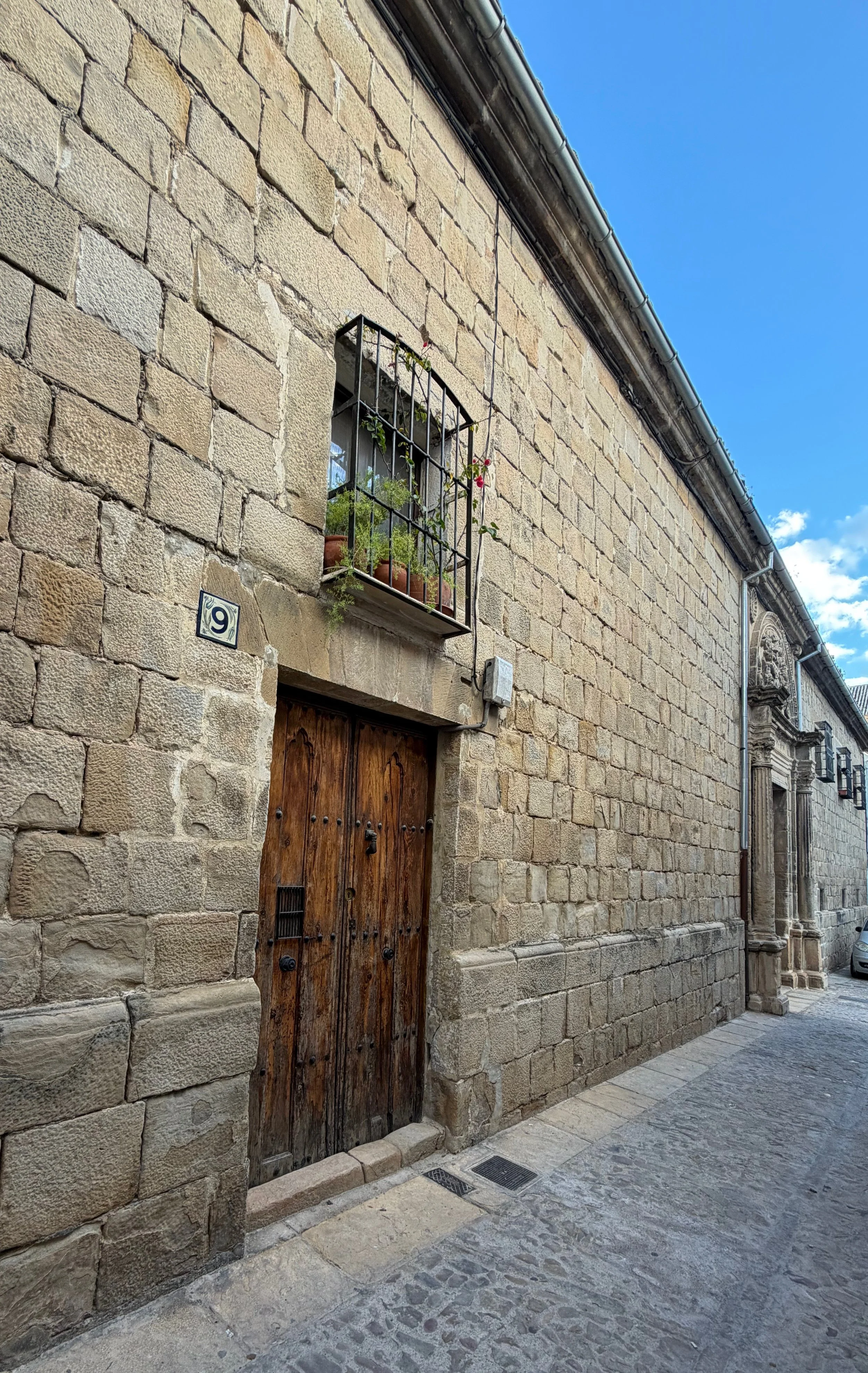 A rustic wooden door set in an old stone wall with an iron-grilled window above it, a small house number sign, and a narrow cobblestone sidewalk. Photo taken Spain, 2025. Doors are a metaphorical for curiosity, being open, and being grounded.