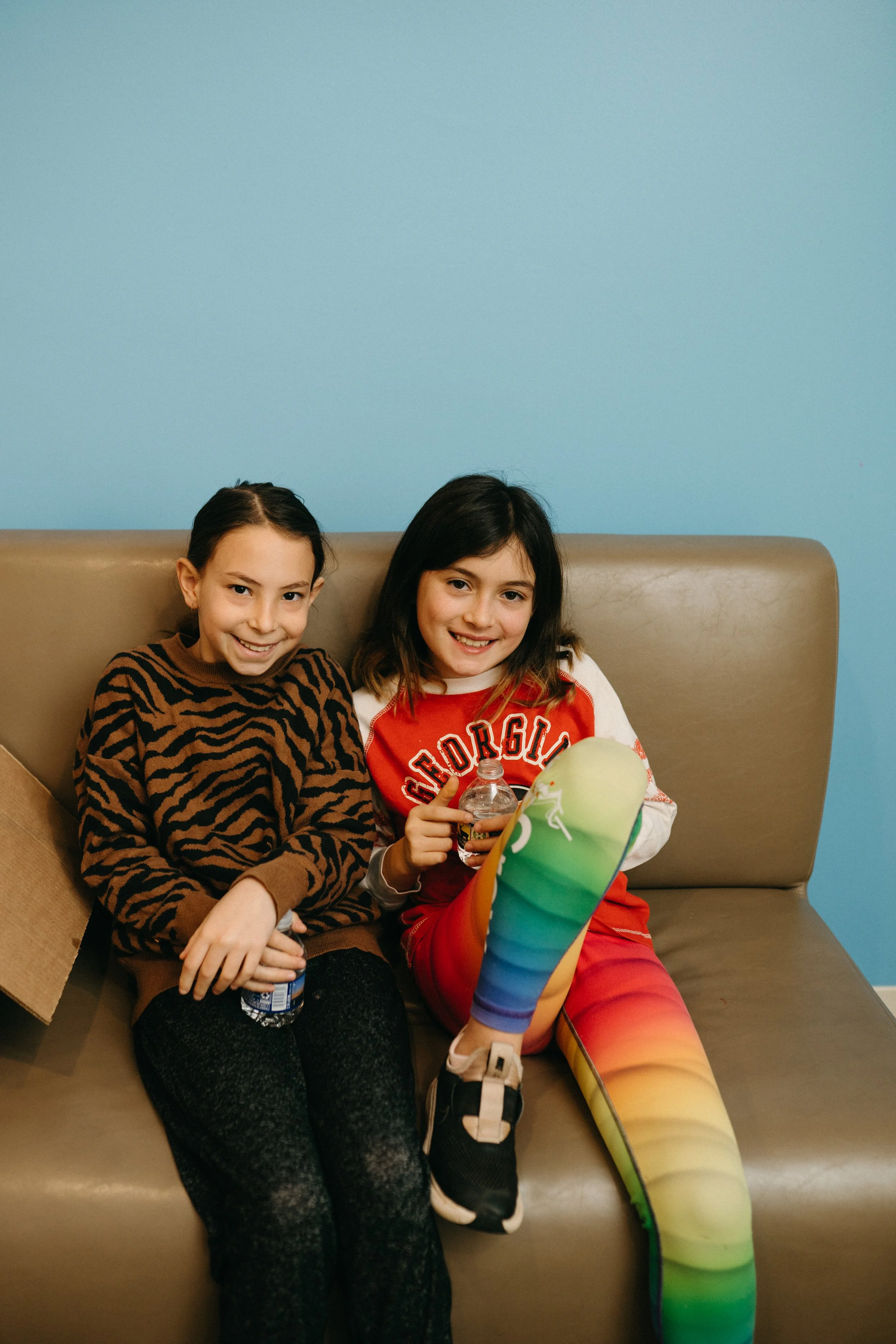 Two young girls sitting on a beige couch, smiling at the camera, with a blue wall behind them. The girl on the left is wearing a tiger-striped sweater and black pants, holding a small water bottle. The girl on the right is wearing a red and white Georgia shirt and rainbow-colored leggings, holding a rainbow-colored inflatable object and a bottle of soda.