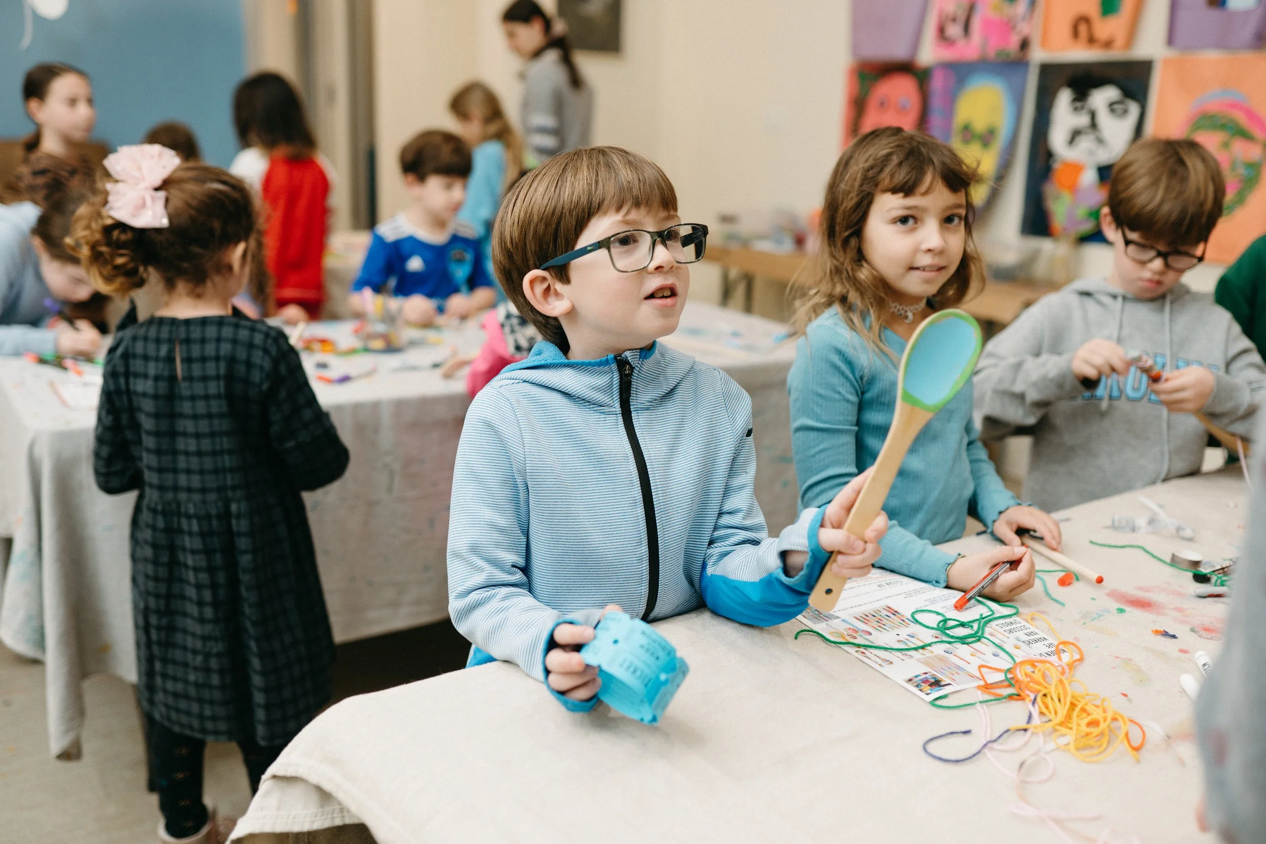 Children at a craft table displaying various materials and artwork in the background in a classroom setting.