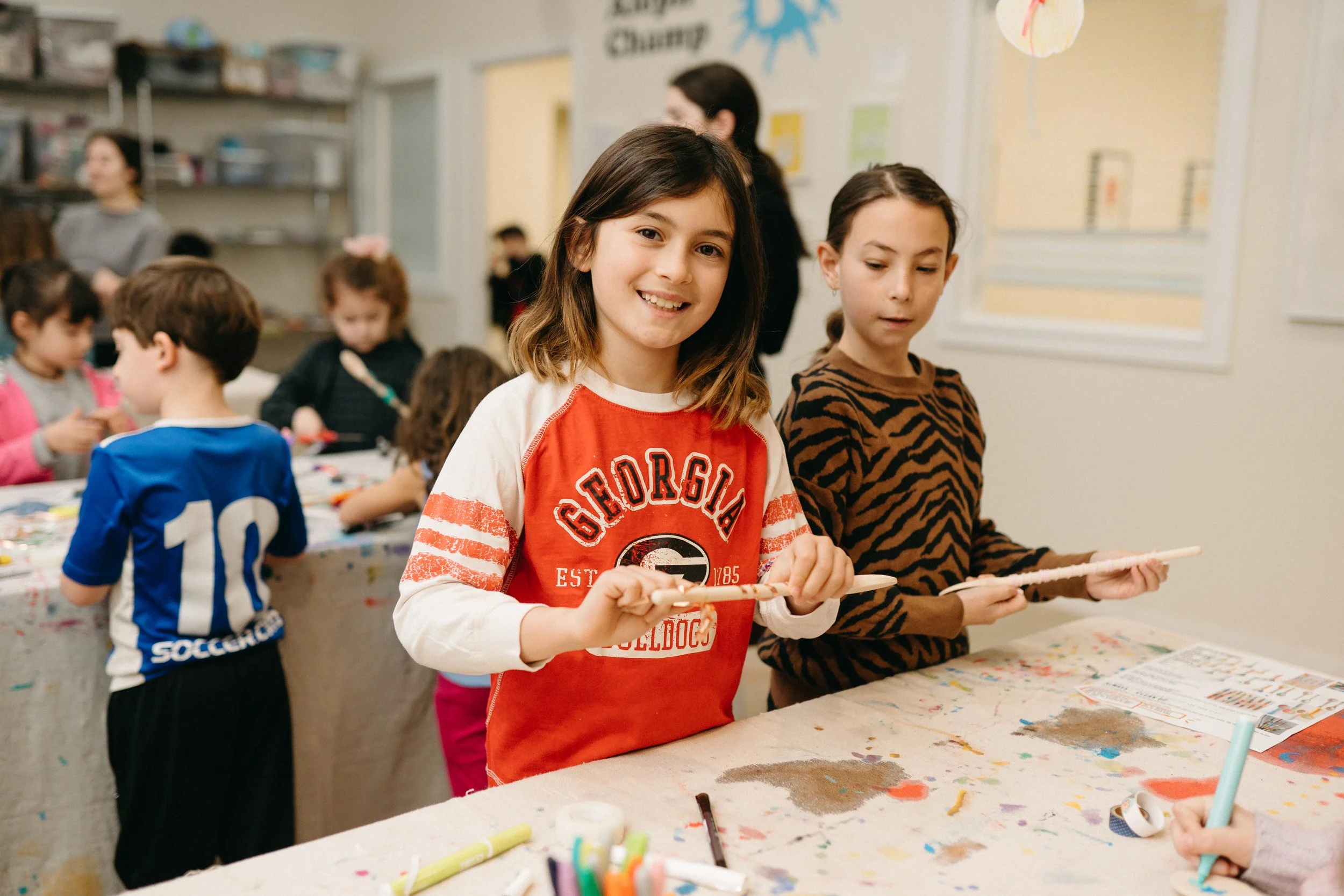 Two young girls participate in a craft activity in a classroom, with other children and teachers in the background. The girl in the foreground wears a red Georgia Bulldogs sweatshirt and holds a stick, smiling at the camera. The girl beside her wears a zebra-striped shirt and looks down at her craft project. The table is covered with art supplies and paper.