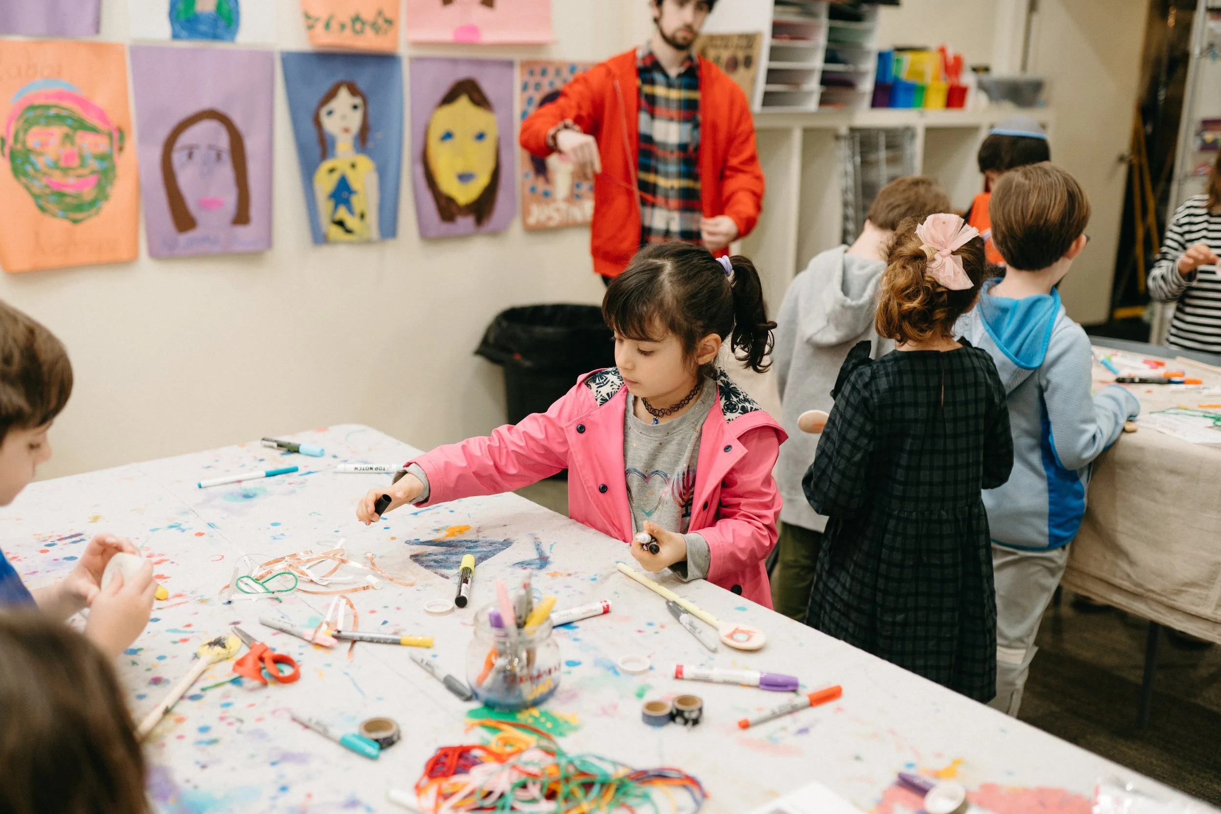 Children and an adult engaged in arts and crafts activities at a long table covered with colorful markers, paper, and ribbon, with children's artwork displayed on the wall behind them.