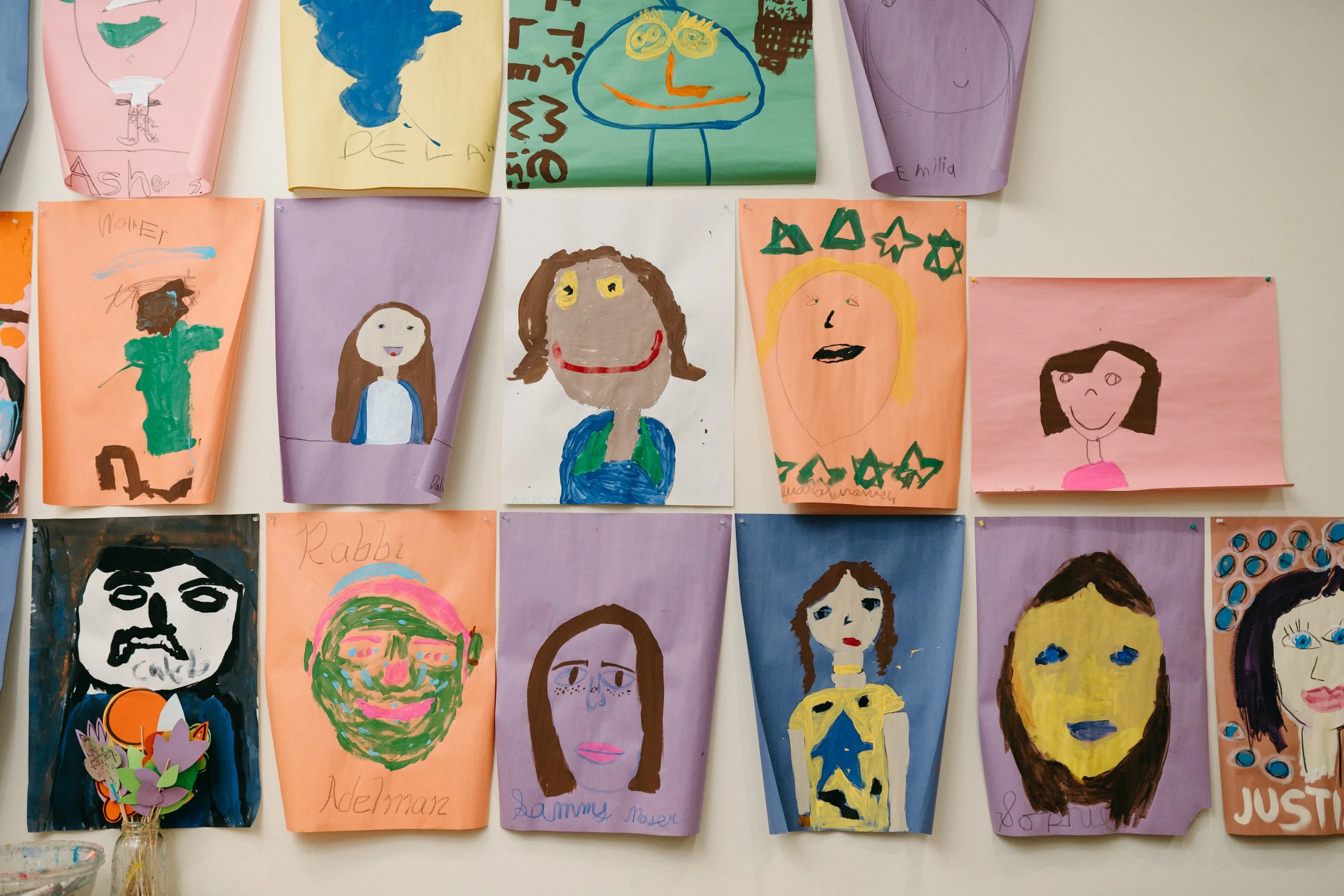 Colorful children's artwork featuring various portraits and faces, displayed on a wall.