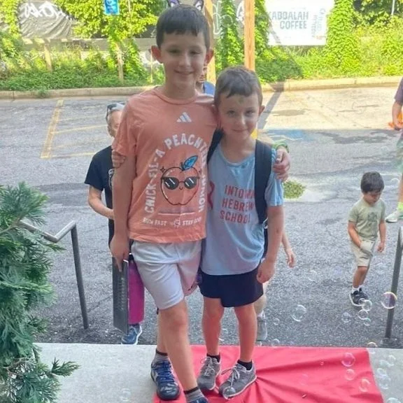 Two boys standing together outside, one taller with a peach T-shirt and the other shorter with a blue T-shirt, surrounded by children and greenery.