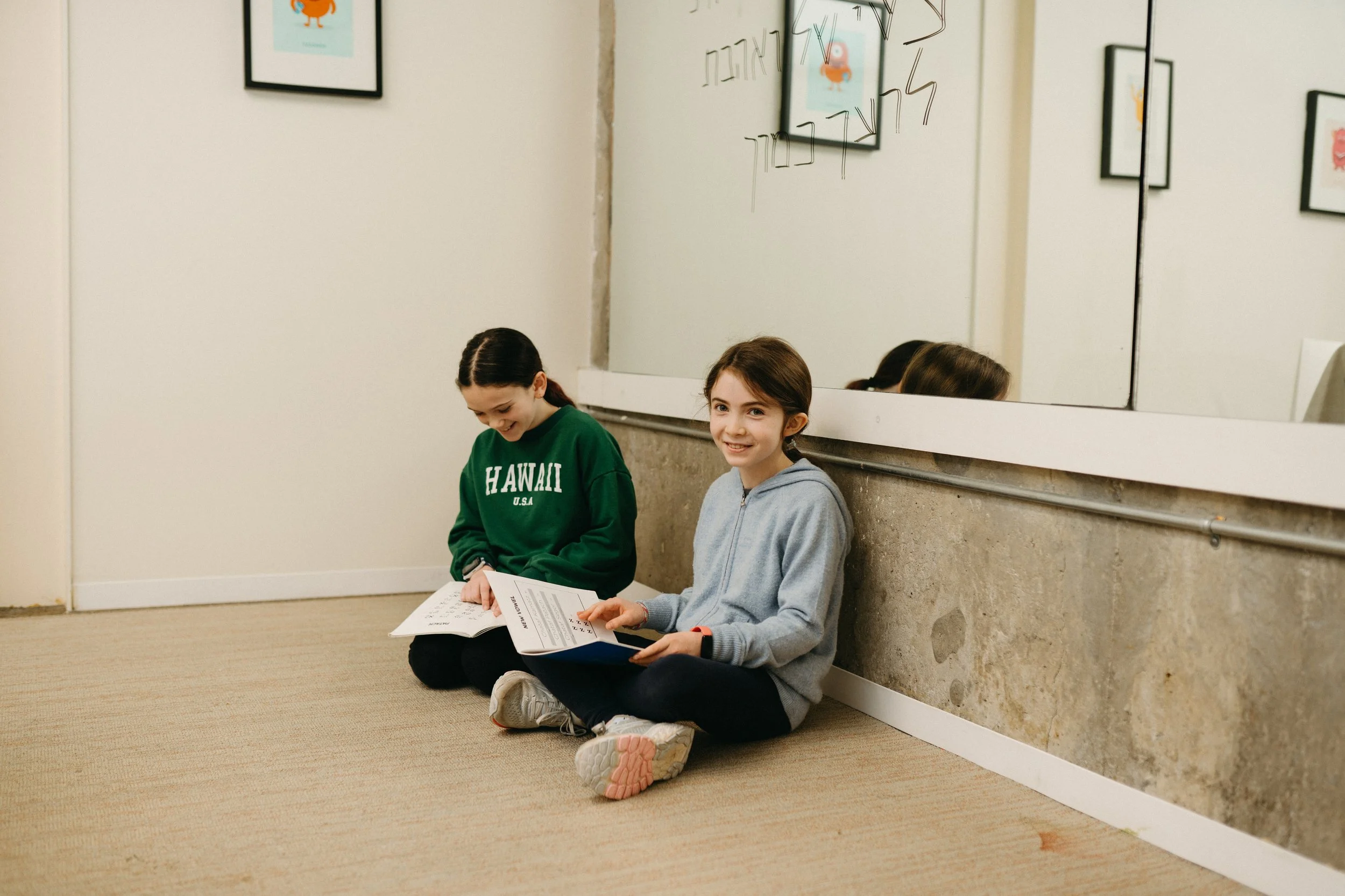 Two girls sitting on the floor against a wall, reading books, with framed pictures hanging above them. One girl is wearing a green Hawaii sweatshirt and the other a gray hoodie, both smiling and engaged with their books.