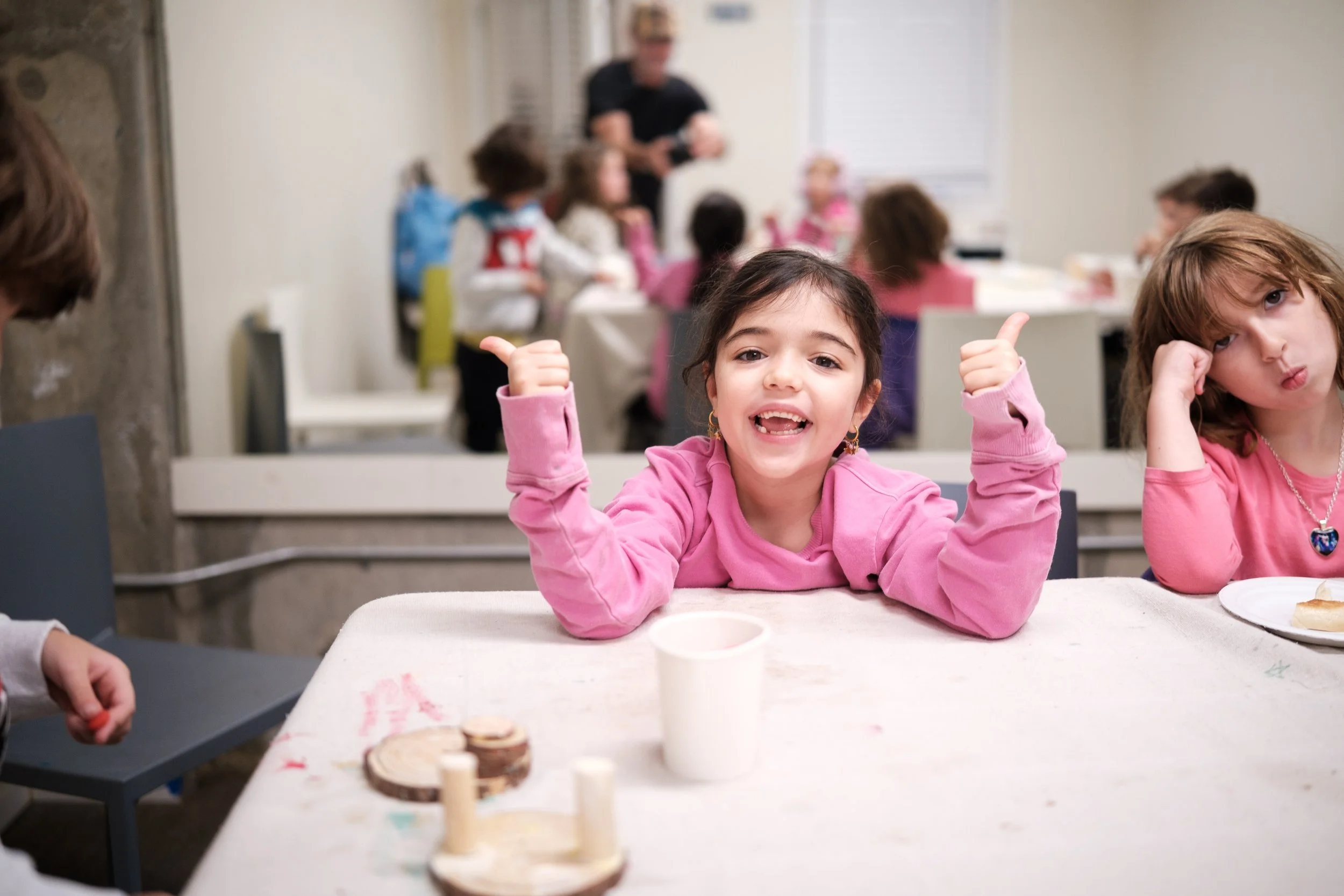 A group of young children sitting at a table in a classroom or activity room, with two girls in pink sweaters smiling and giving thumbs-up, while other children are blurred in the background.