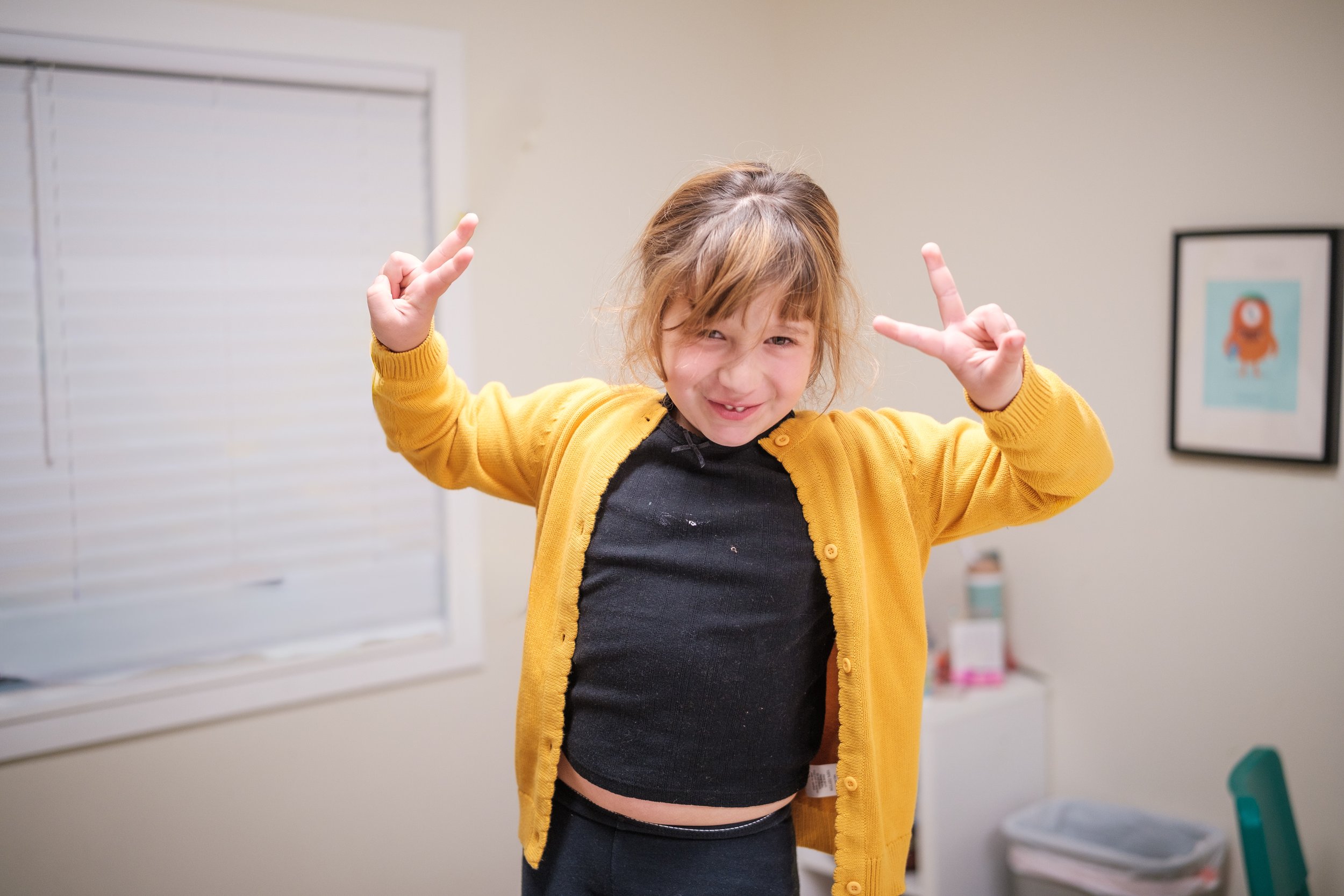A young girl with short brown hair, wearing a yellow cardigan and a black shirt, is making a peace sign with both hands and smiling mischievously in a room with white walls and a framed picture in the background.