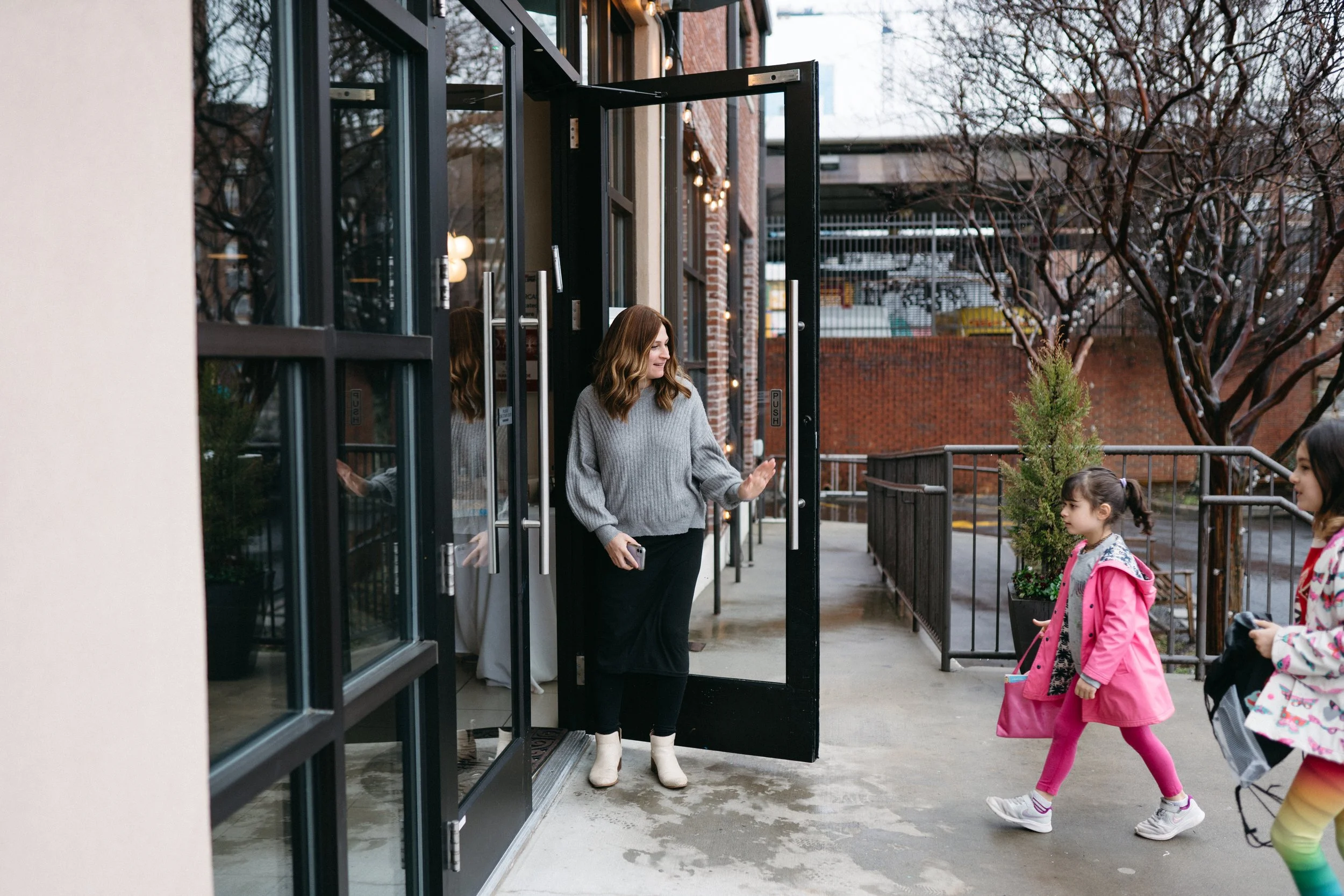 A woman with wavy brown hair, wearing a gray sweater, black pants, and white boots, is standing at an open door of a building, smiling and waving to children outside. There are three children in colorful clothes walking past, one in a pink jacket and another in a rainbow-colored jacket. The building has brick and cream-colored walls, and the scene is outdoors with leafless trees and a parking garage in the background.