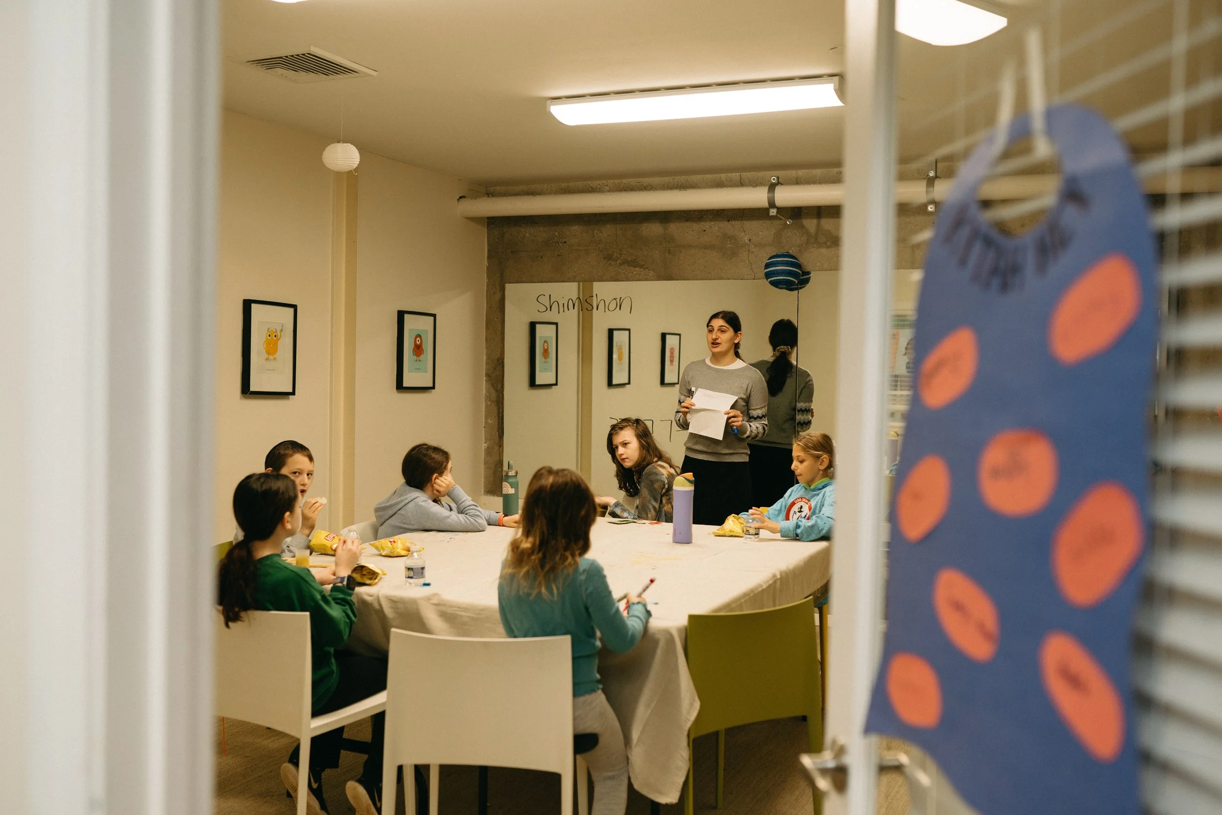 A group of children sitting around a table with snacks, listening to a woman standing and speaking to them in a room decorated with framed artwork and hanging paper lanterns, with a sign that says 'Shimshon' on the wall.