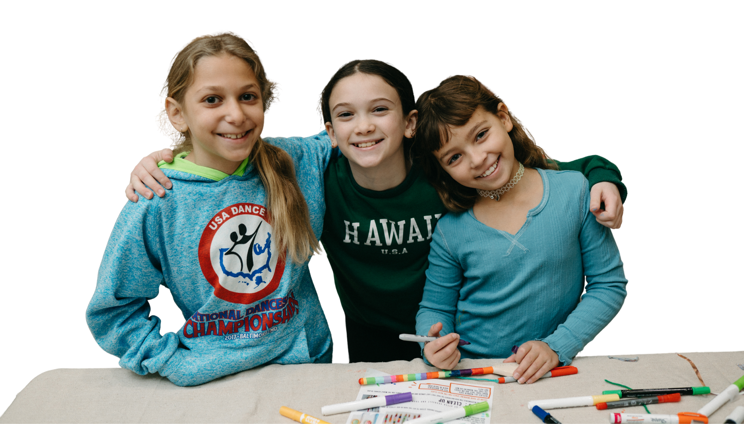 Three smiling young girls hugging each other, with markers and paper on a table in front of them.