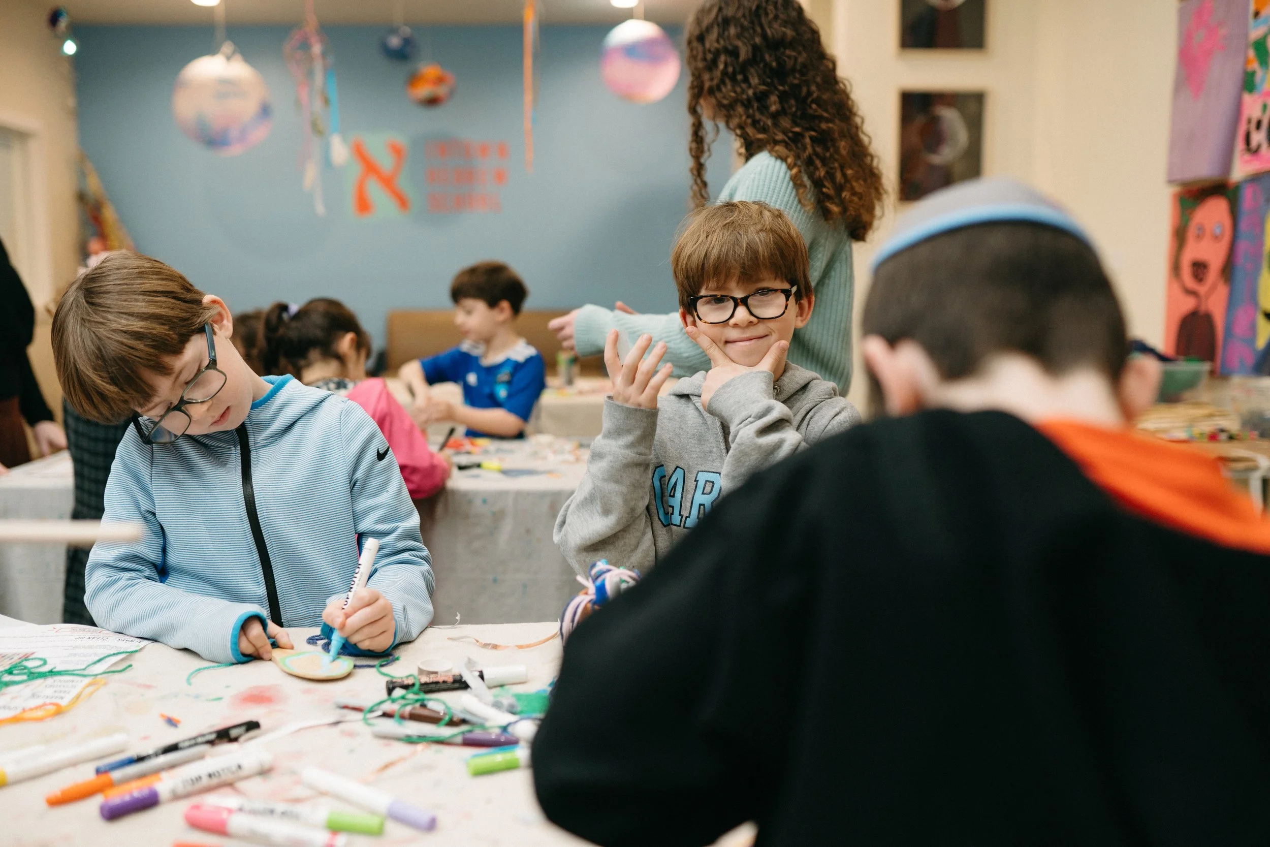 Children engaging in arts and crafts at a table, with markers and craft supplies, in a classroom decorated with colorful artwork and hanging decorations.