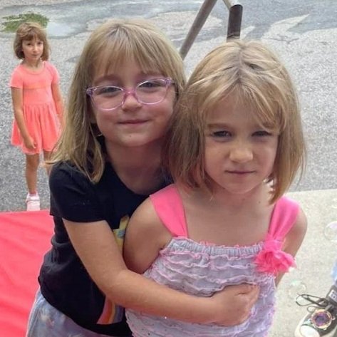 Two young girls hugging outdoors, with a third girl standing in the background, on a pink blanket near water.