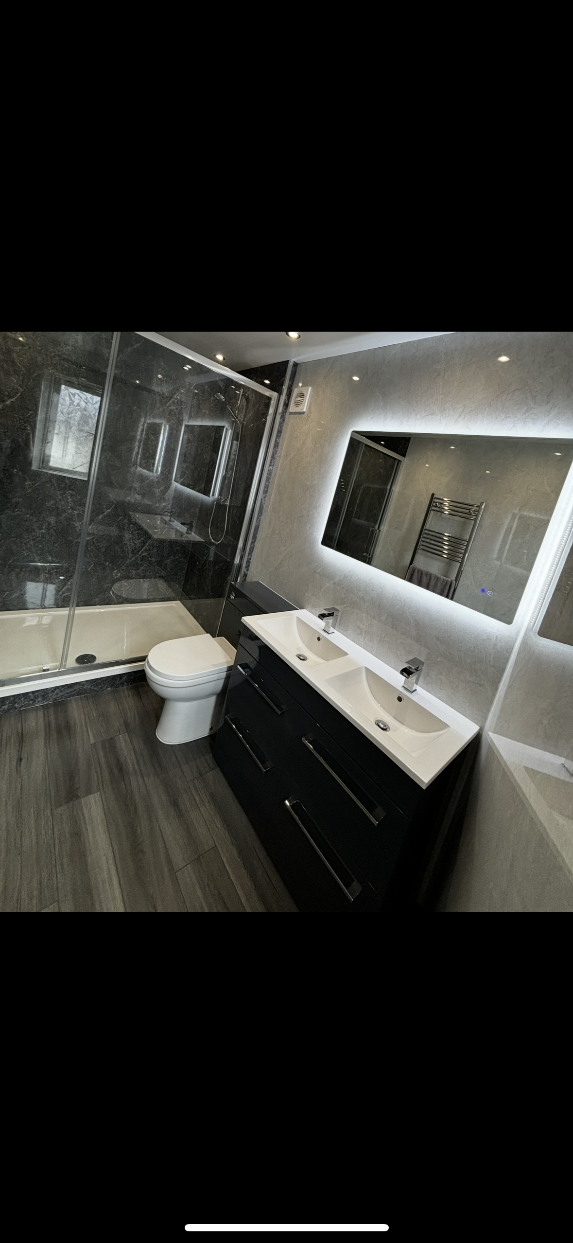 Modern bathroom with dual sinks, a large illuminated mirror, a black vanity, a white toilet, and a glass-enclosed shower with black marble walls.