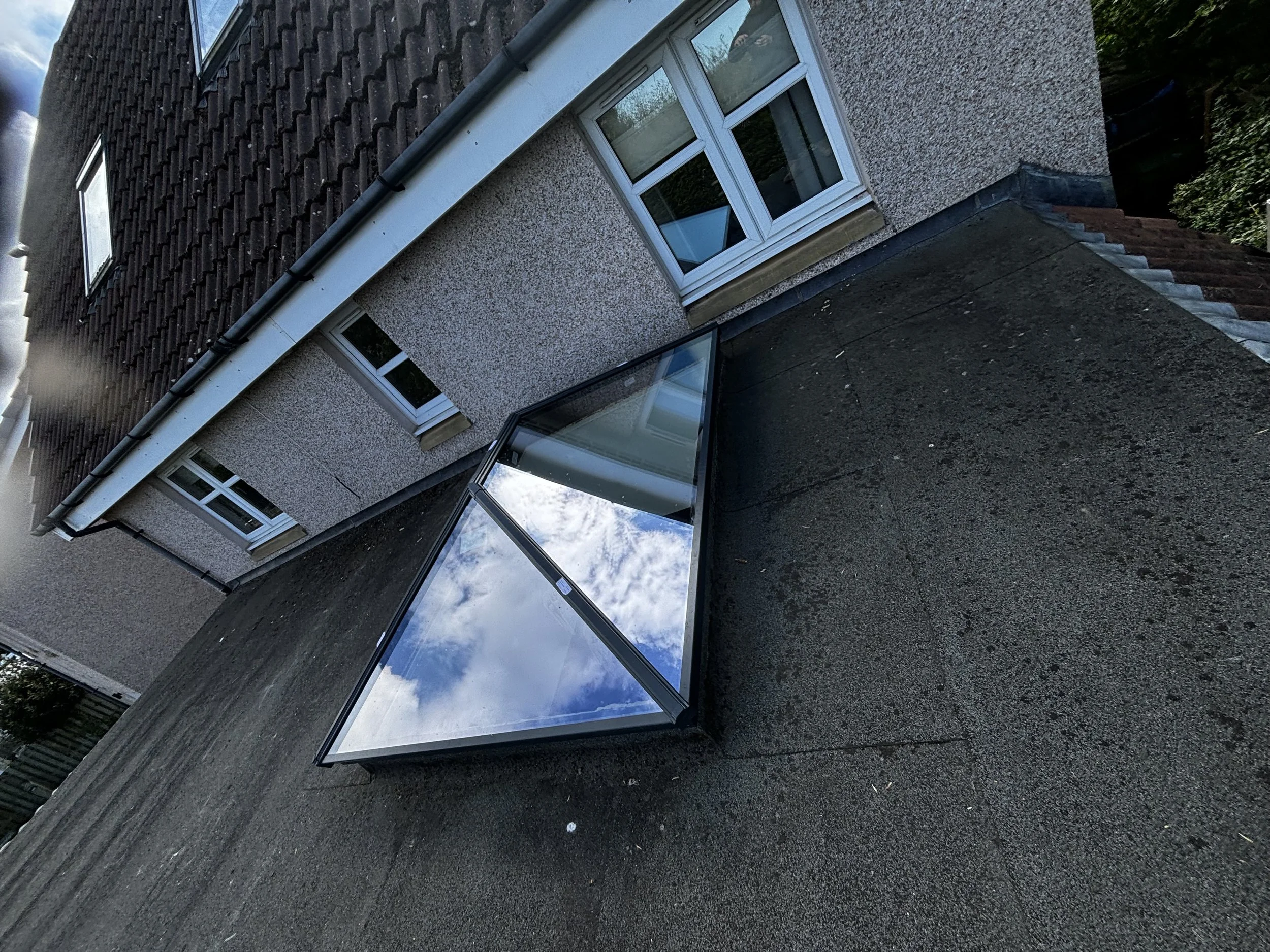 Flat rooftop with a glass structure reflecting the sky and clouds, adjacent to a residential building with windows, textured walls, and a sloped roof.