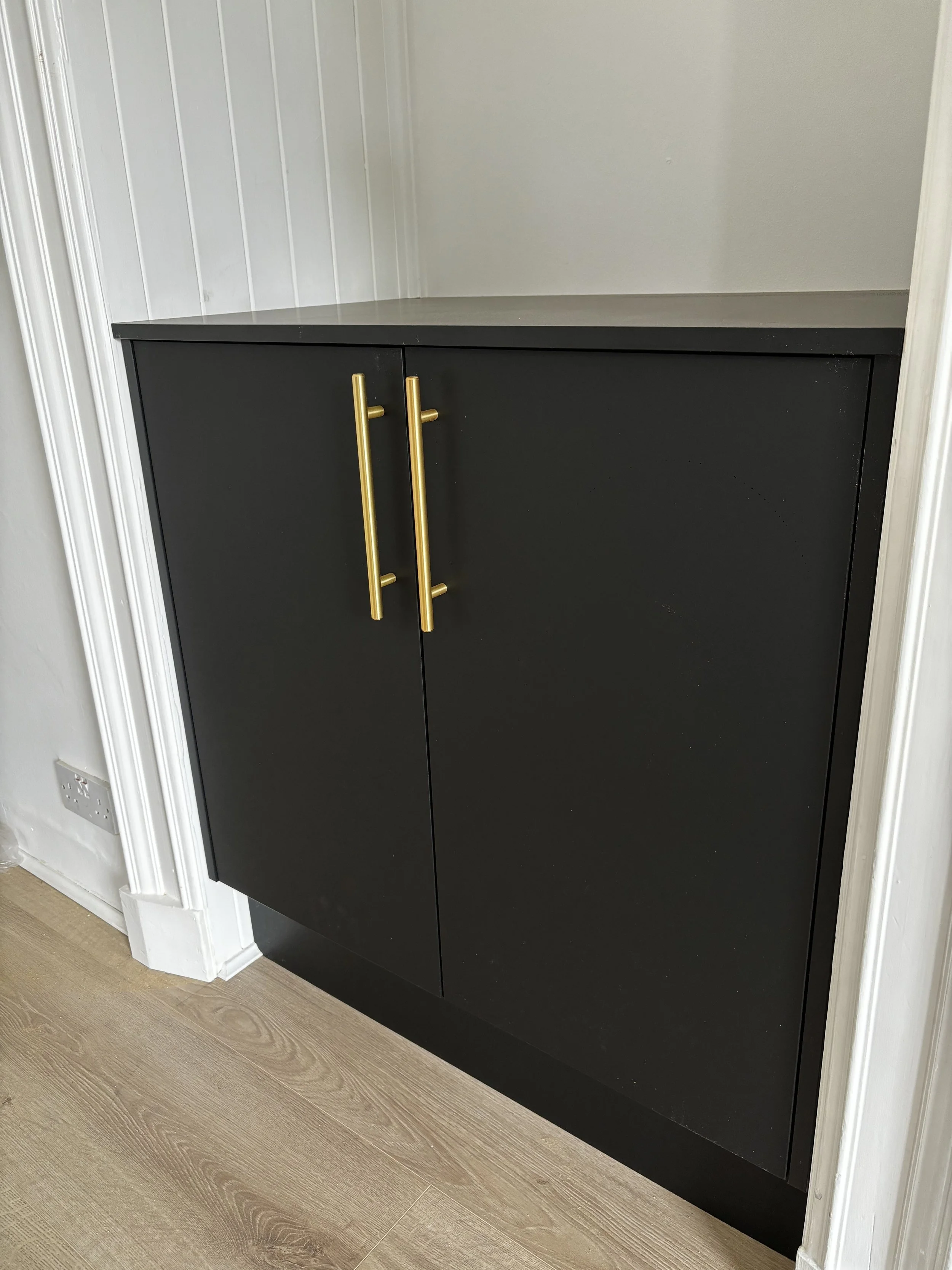 A black cabinet with gold handles in a white room with wood flooring.