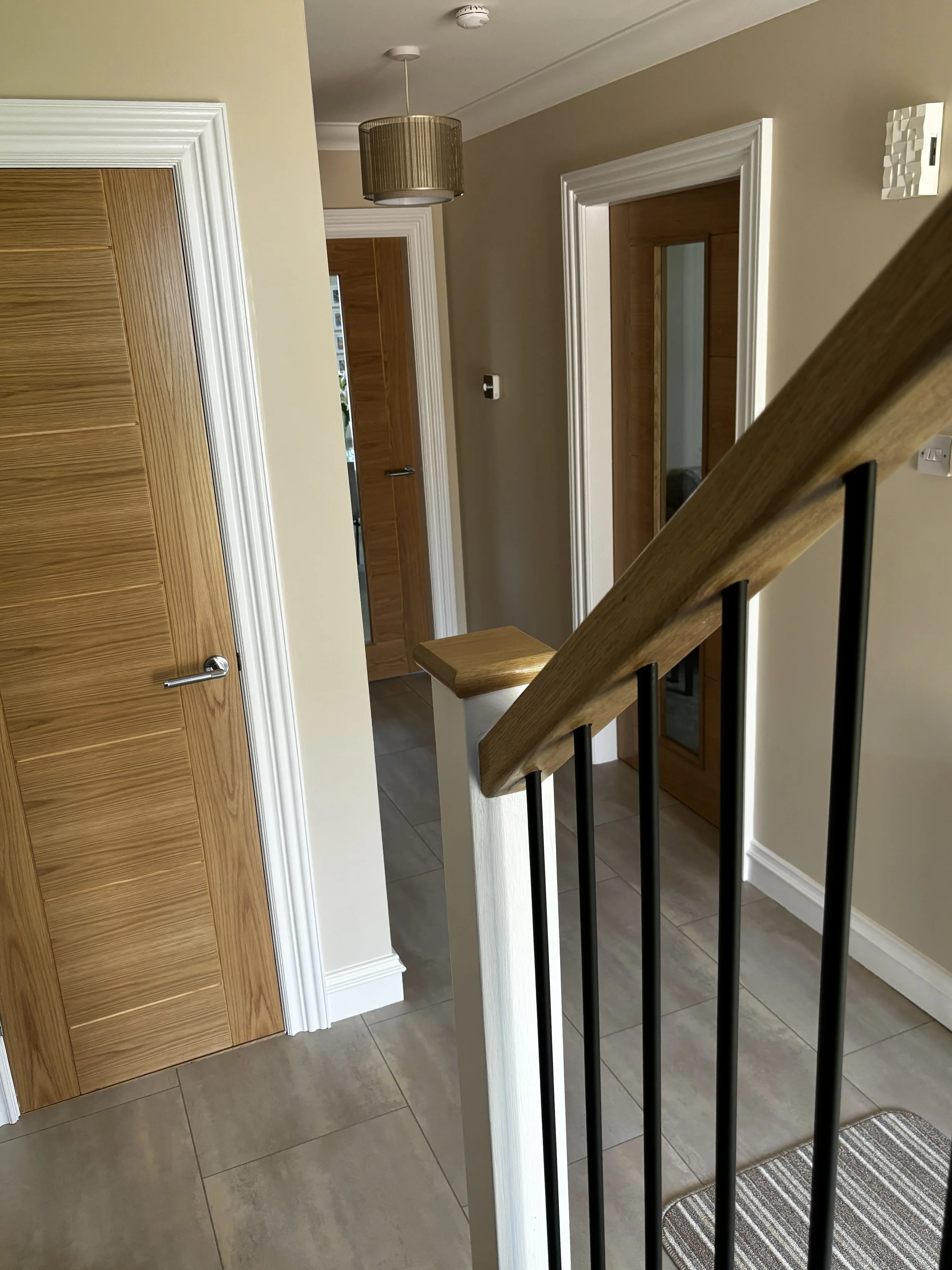 Interior view of a hallway with beige walls, wooden doors, white trim, a staircase with a wood and black metal handrail, tiled floor, a ceiling light fixture, and a striped rug.