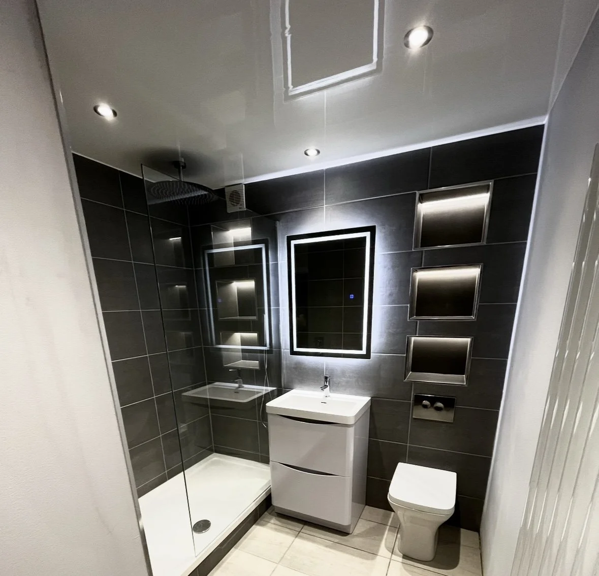 Modern bathroom with dark tiled walls, a walk-in shower with glass door, illuminated mirror above a small white vanity, and a wall-mounted toilet with built-in shelves.