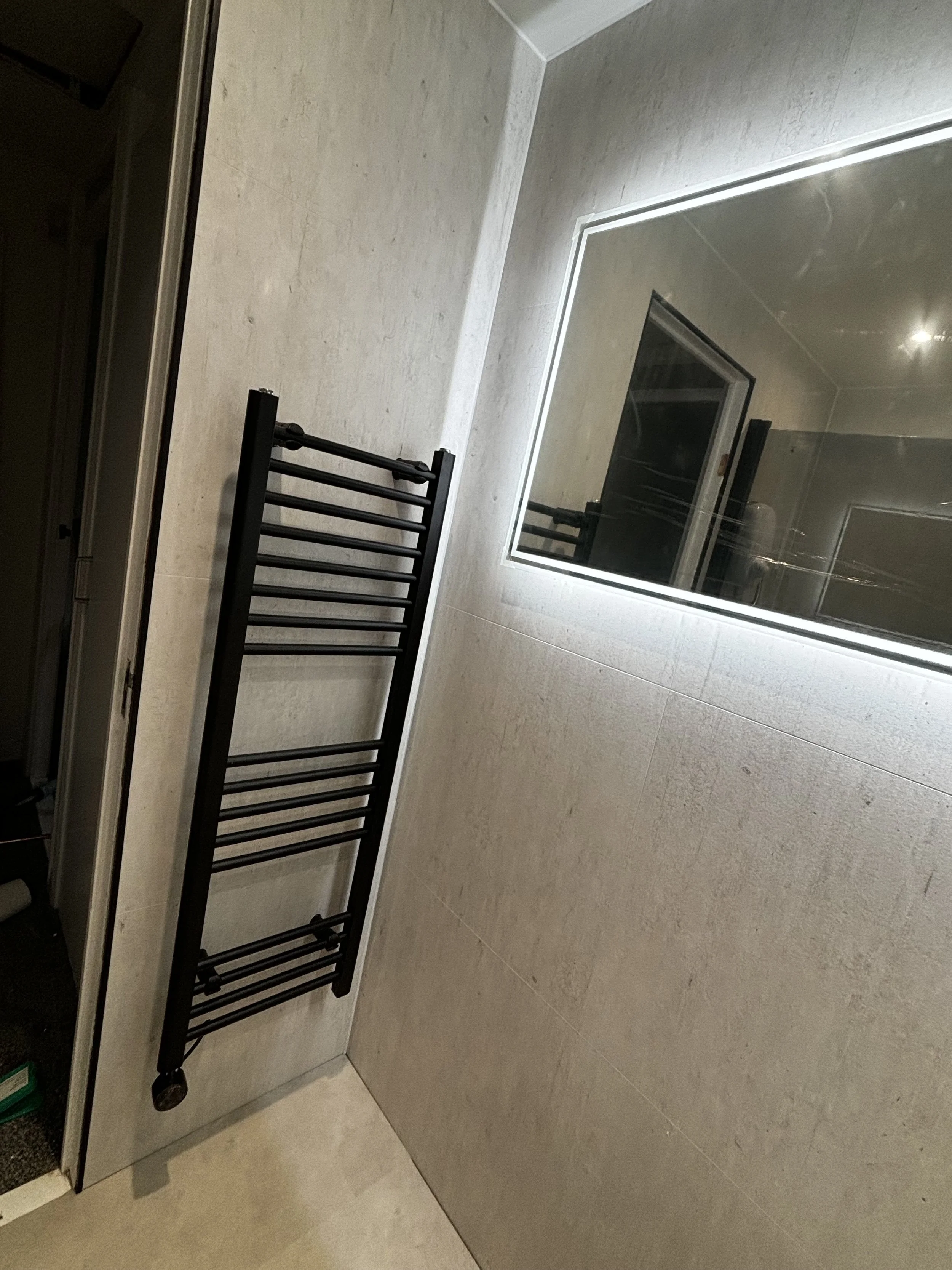 A bathroom with a black towel warmer mounted on a beige tiled wall, and a large mirror reflecting the space.