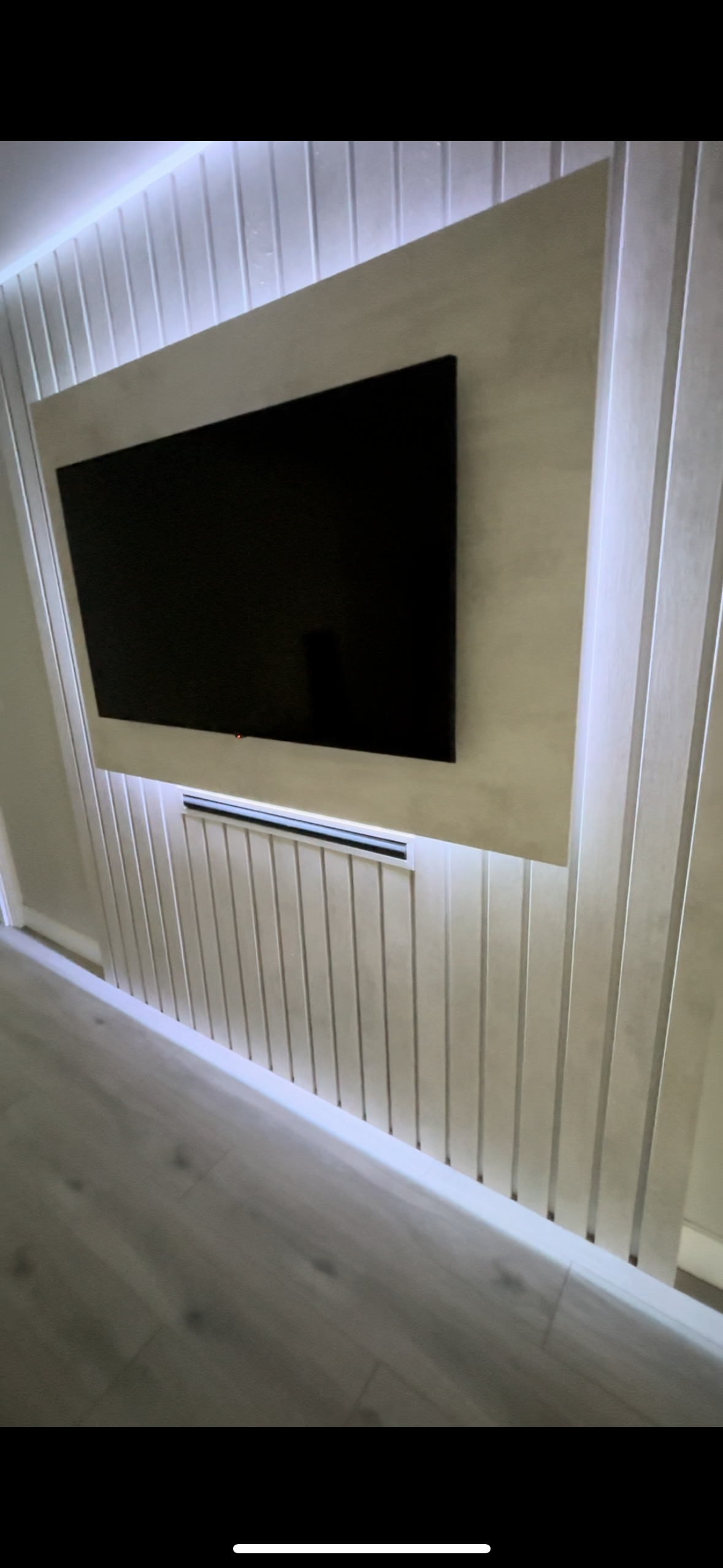 A flat-screen television mounted on a beige wall panel, with a white wall and wood-paneled wall behind it.