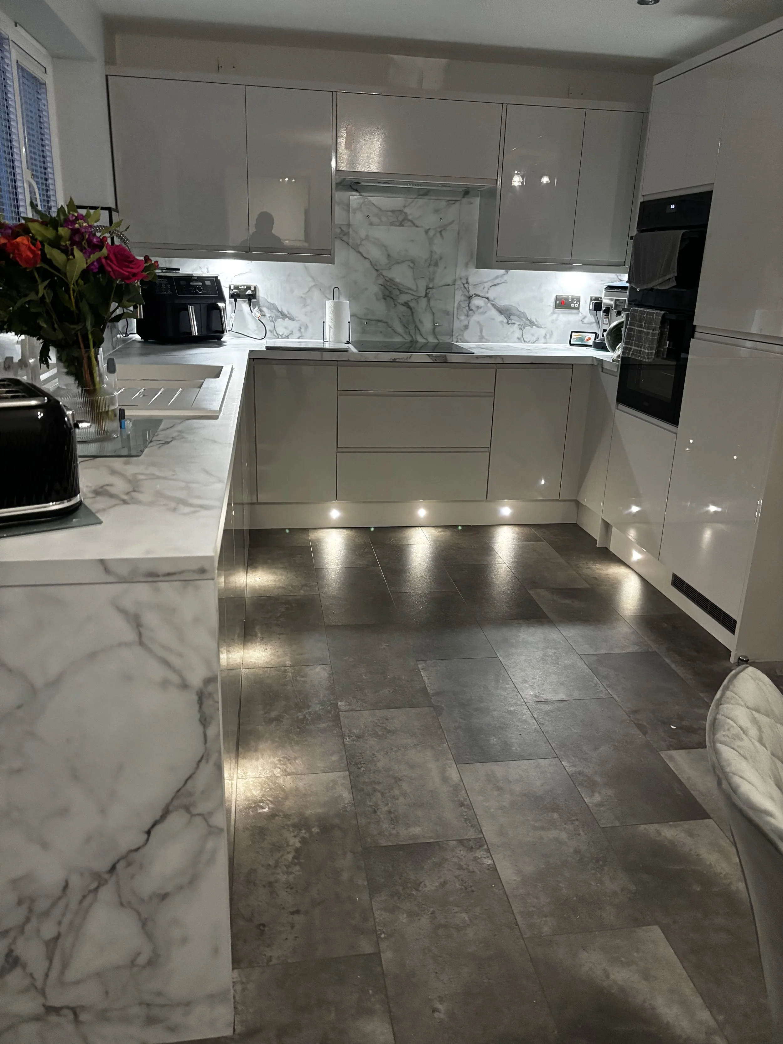 Modern kitchen with white marble countertops and backsplash, gray cabinets, and dark tile flooring. Contains a black toaster, a bouquet of colorful flowers, and kitchen appliances.