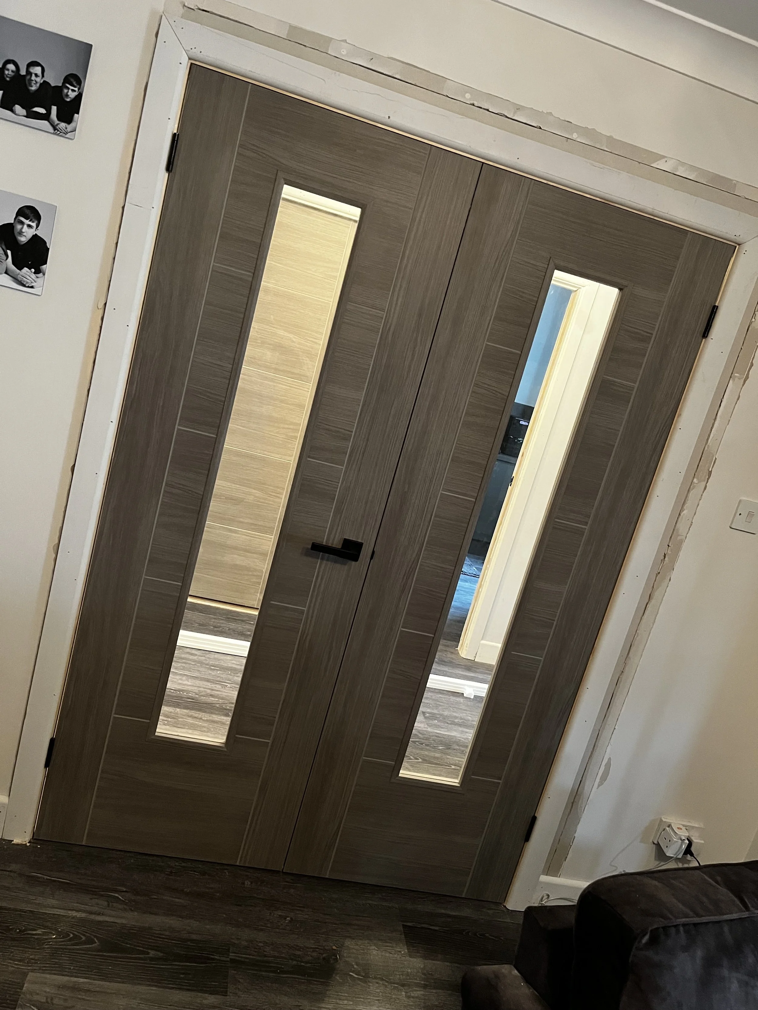 A double wooden door with glass panels, closed, in a room with dark wood flooring and partially finished white walls. Parts of the wall around the door are unfinished, and there are two black-and-white photos of people on the left side of the image.