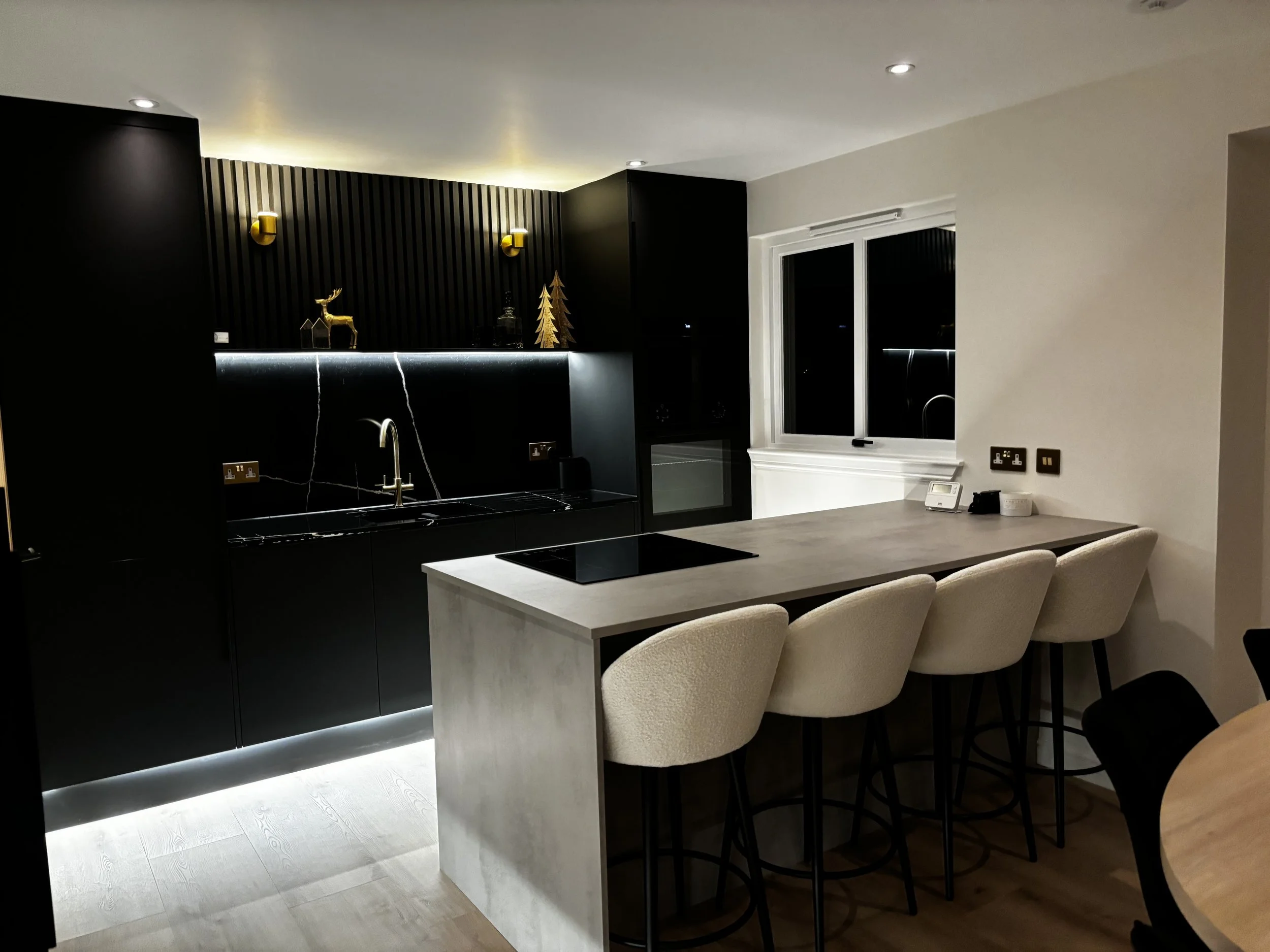Modern kitchen with black cabinetry, a light-colored countertop island with four cream upholstered bar stools. Decor includes gold accents, a gold reindeer and a gold Christmas tree on a shelf, and two gold wall sconces. There is a window with black 